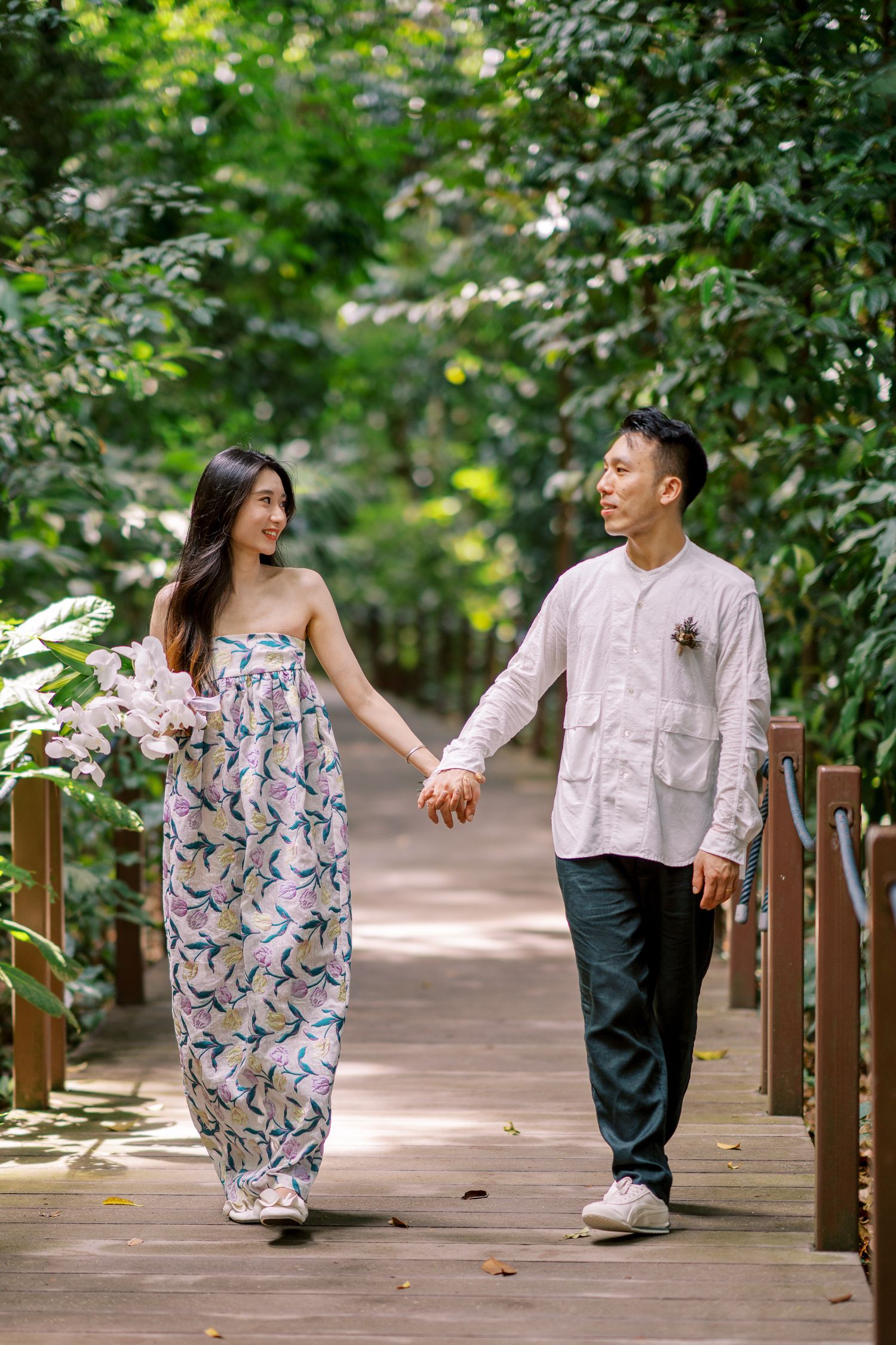 Playful couple in coordinated outfits dance along a wooden boardwalk in a tropical garden setting.