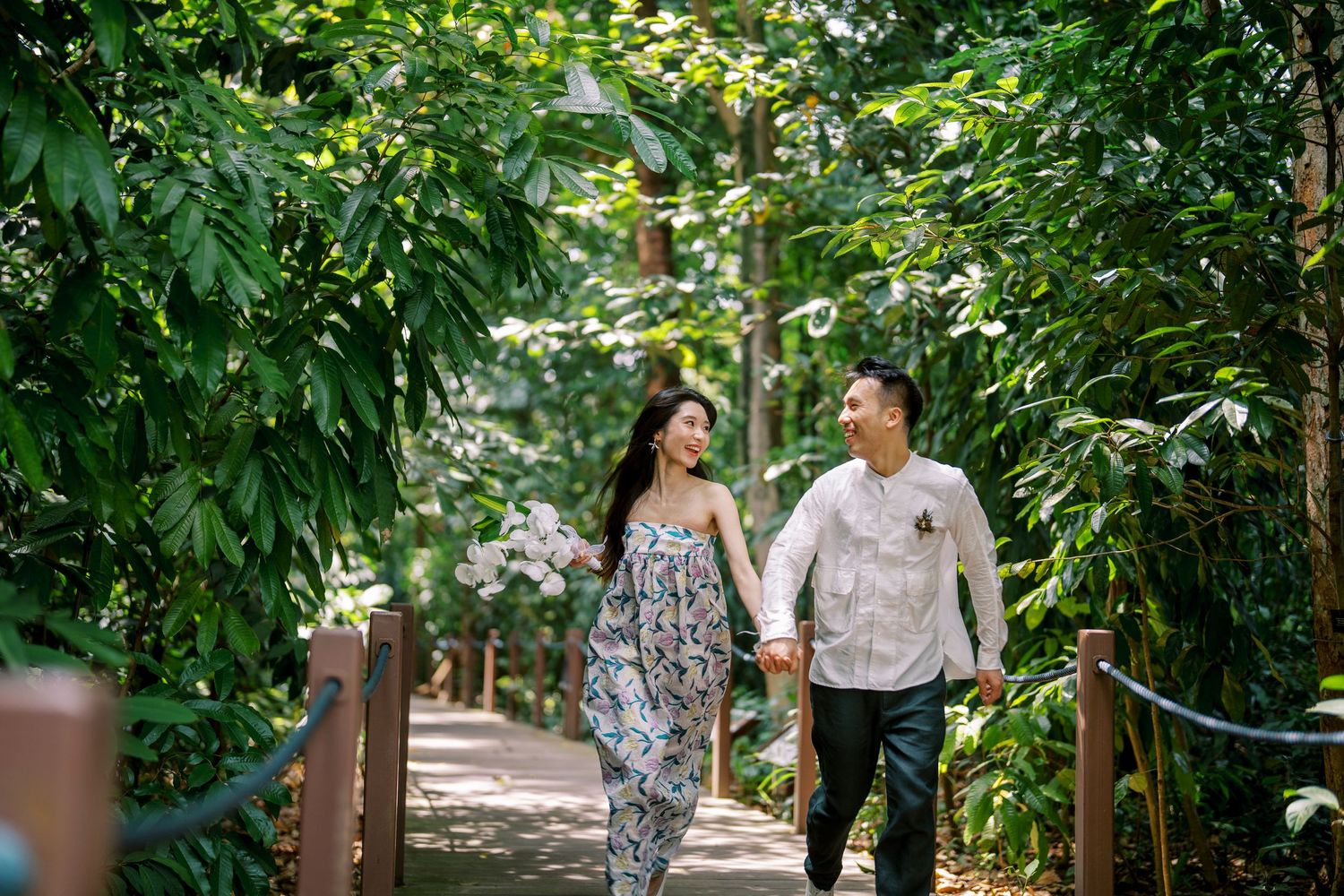 A couple strolls hand in hand through a sunlit garden path with wooden railings.