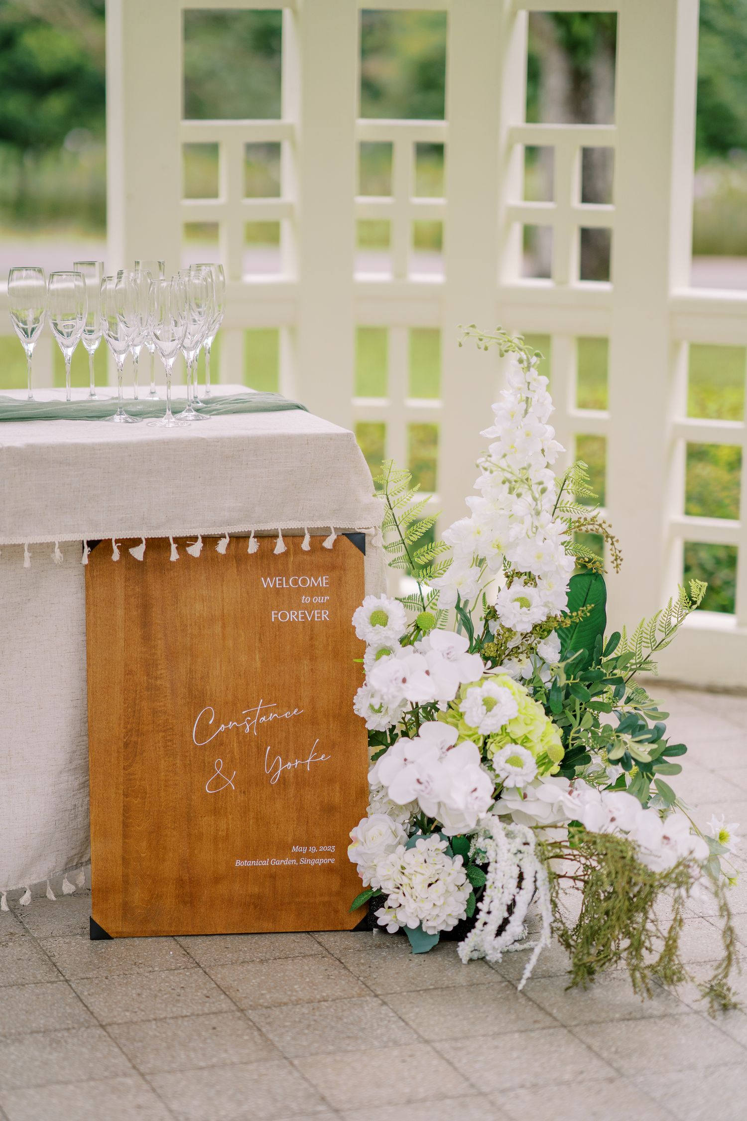 Elegant wedding welcome sign with wooden board and white floral arrangement on a table with champagne glasses.