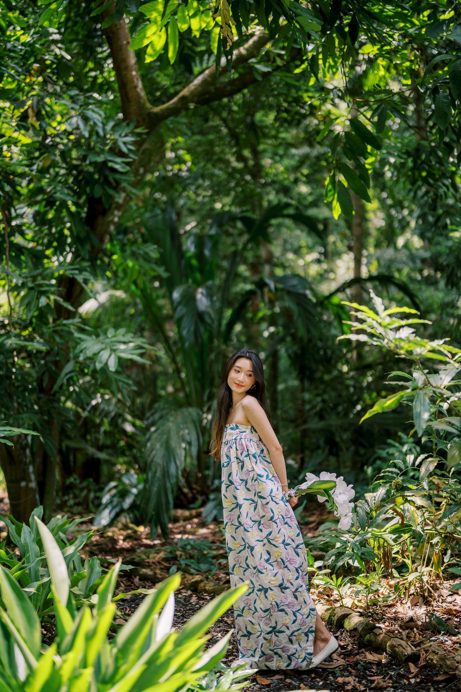Woman in a floral dress poses gracefully in a lush tropical garden setting.