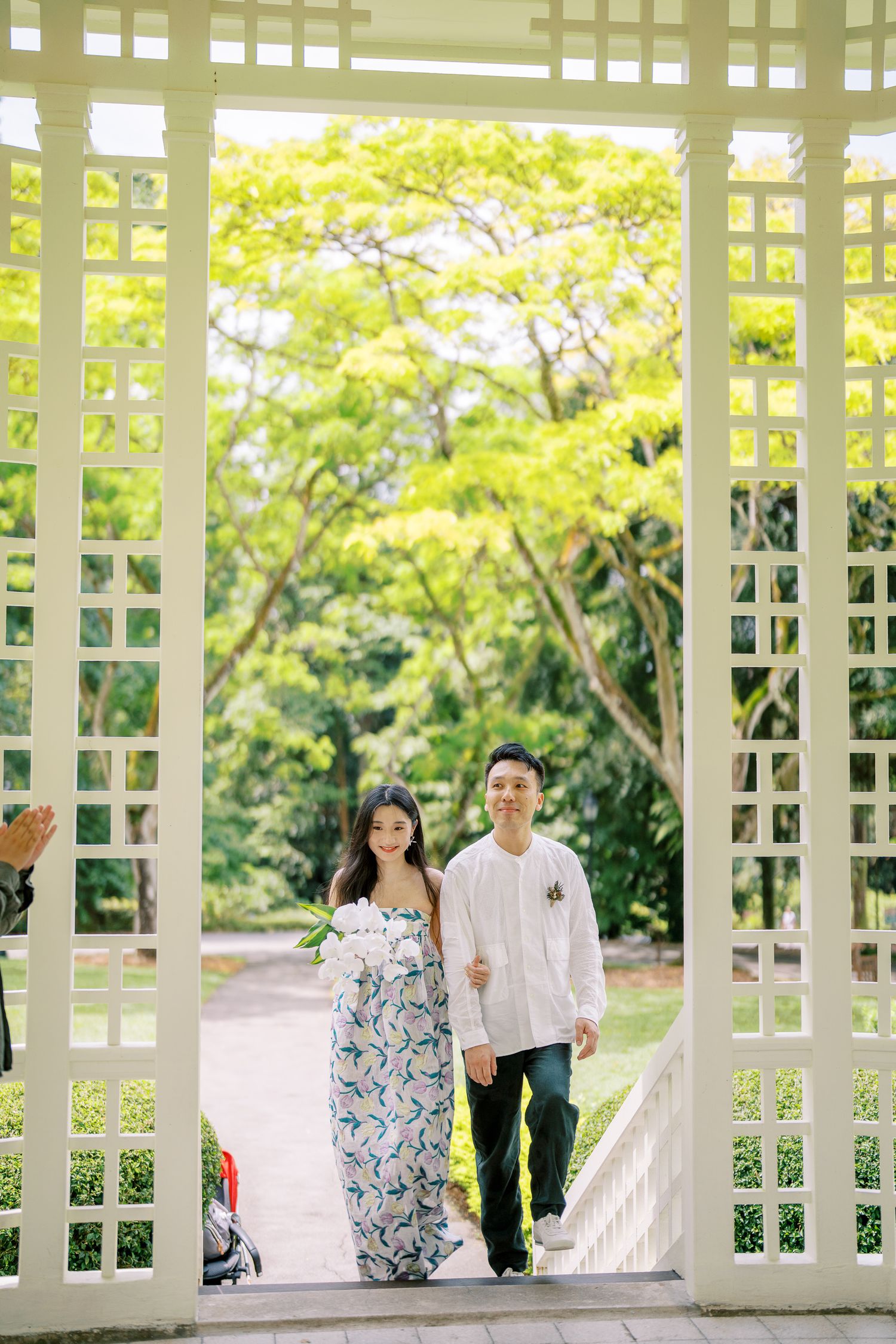 A couple strolls together through an elegant white latticed pavilion in a garden setting.