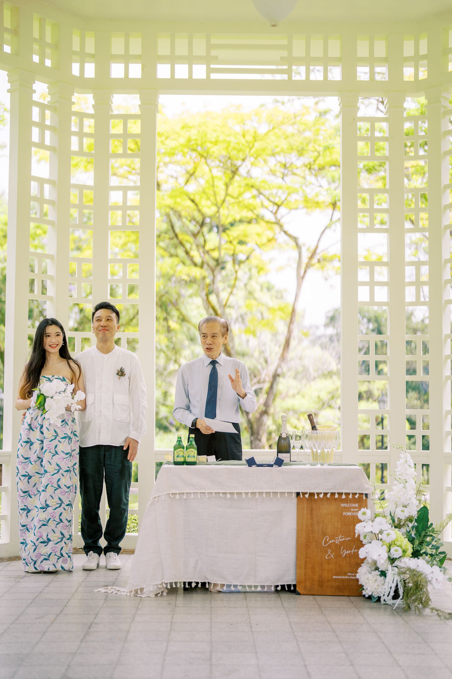 An outdoor event setup under a white latticed structure with tables and flowers.