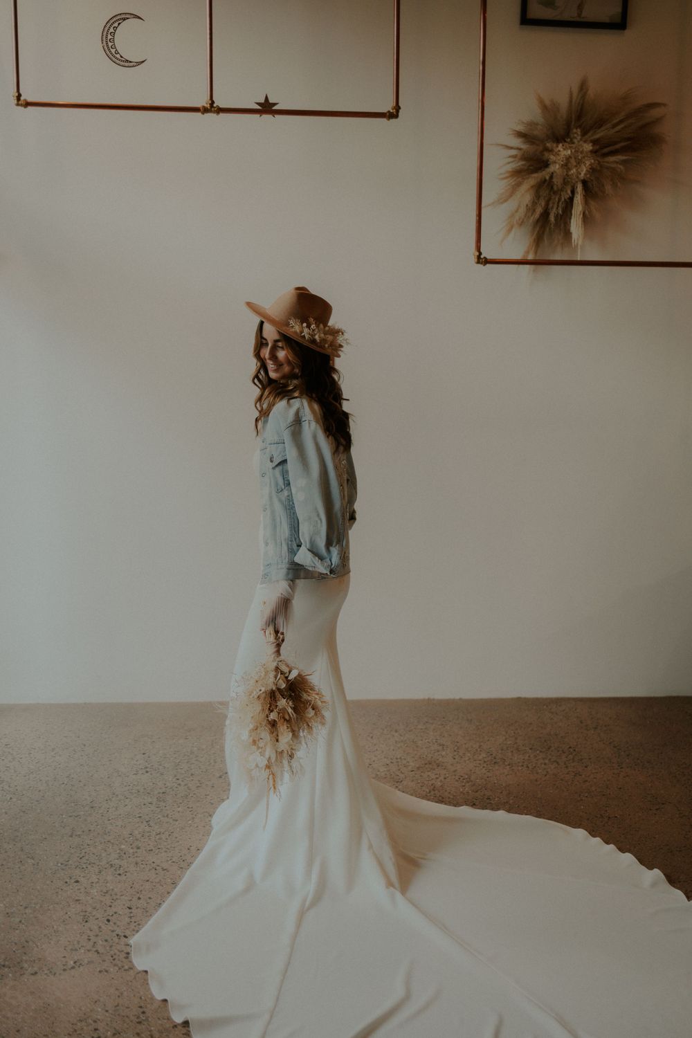 Wedding dress with denim jacket and felt hat creating a modern boho bridal look.