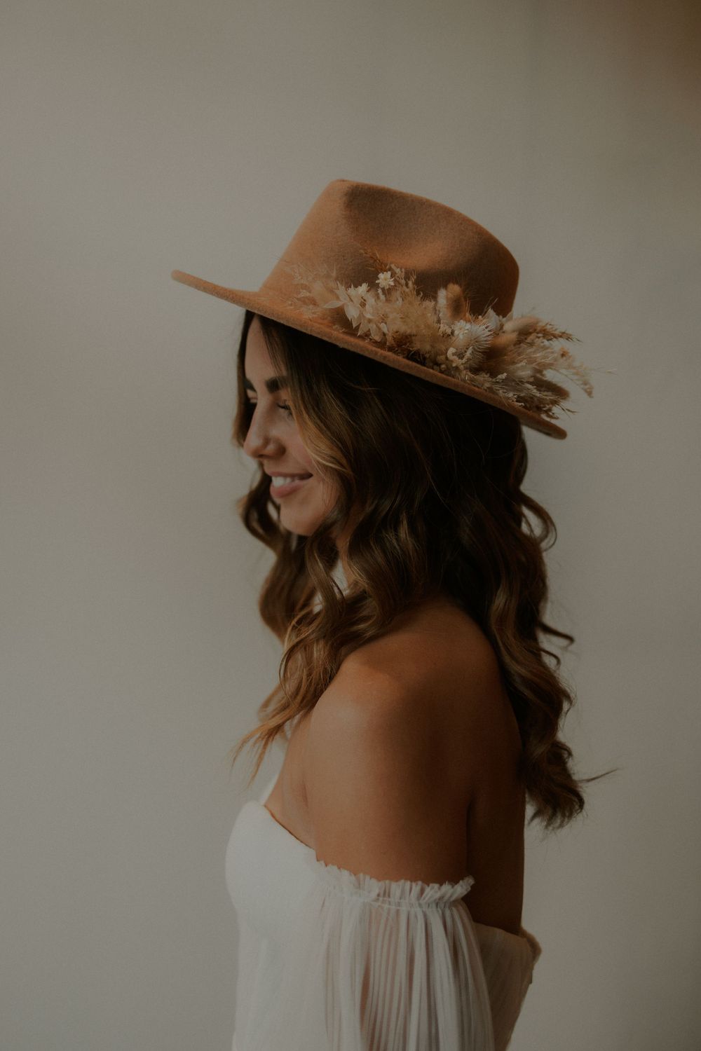 A stylish tan felt hat with dried flowers adorns a white off-shoulder dress in a moody portrait.