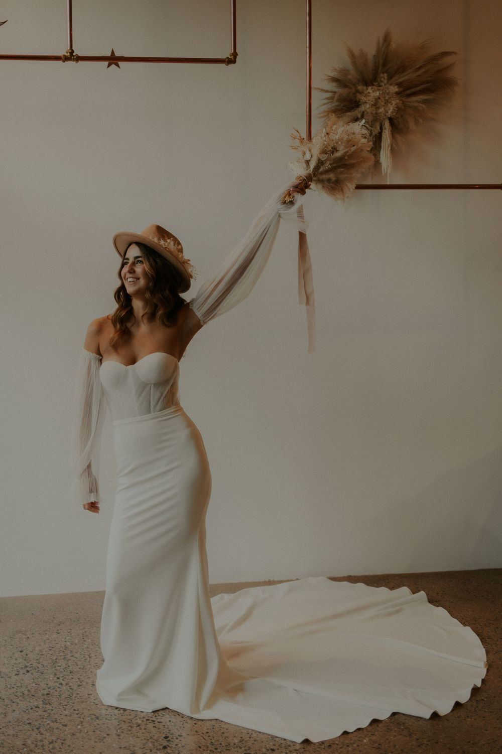 Stylish bride in fitted white gown with brown hat poses against minimalist wall with dried florals.