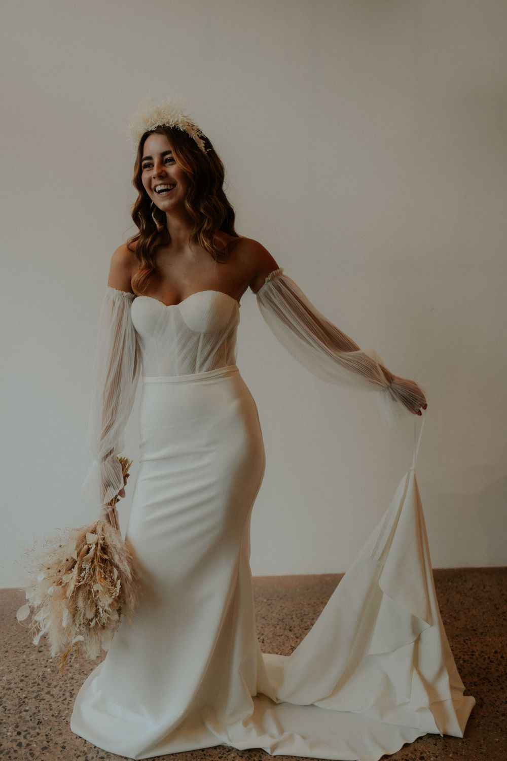 Elegant bride in form-fitting white gown with sheer sleeves holding dried floral bouquet.