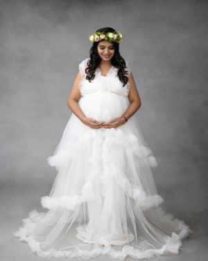 Maternity portrait in flowing white dress with floral crown against grey background showing pregnancy glow.