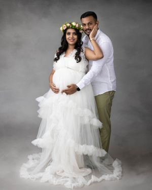 Romantic maternity portrait of couple in white attire with floral crown against gray backdrop.