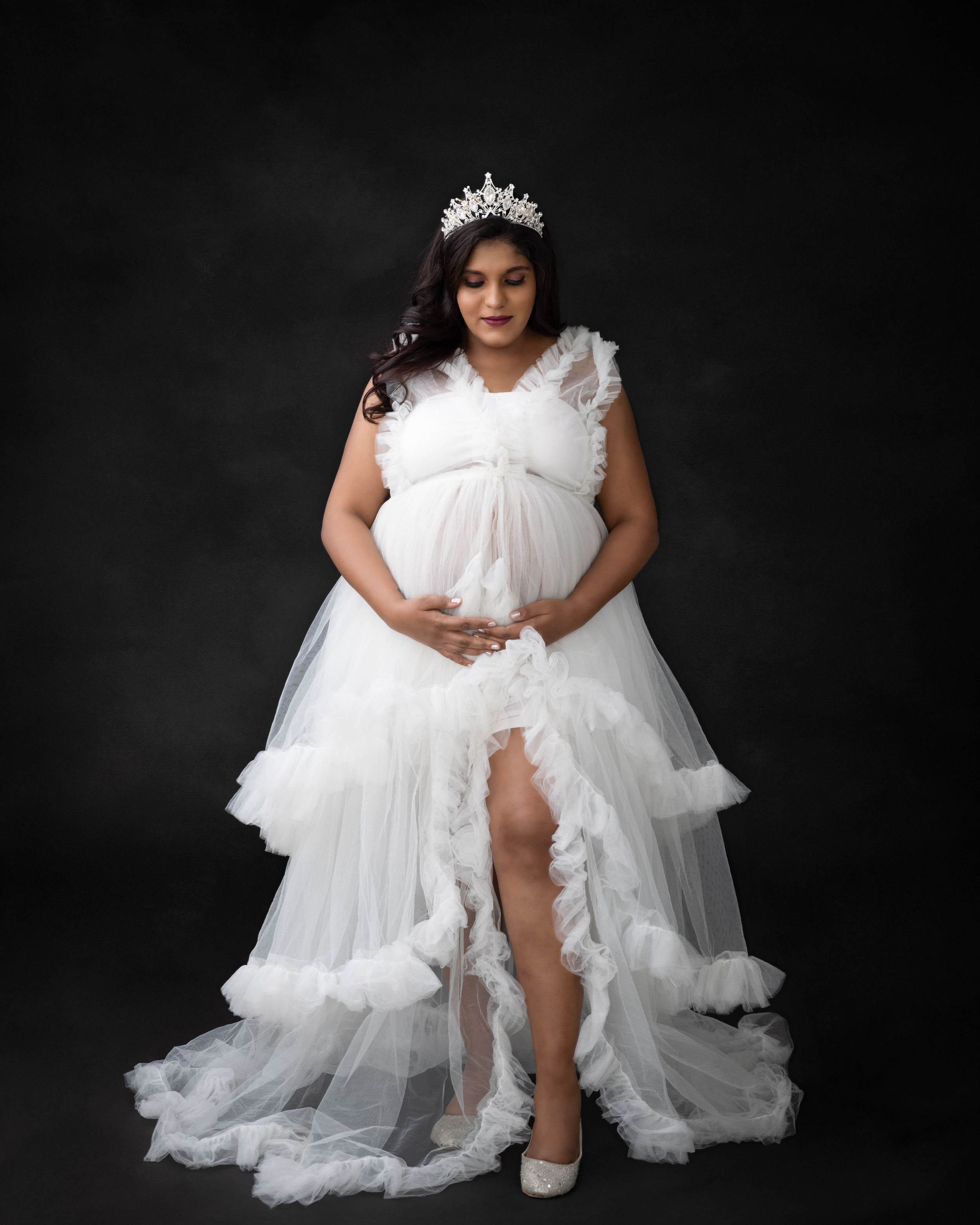 Elegant maternity photo in white tiered gown with crown accessory against dark backdrop.
