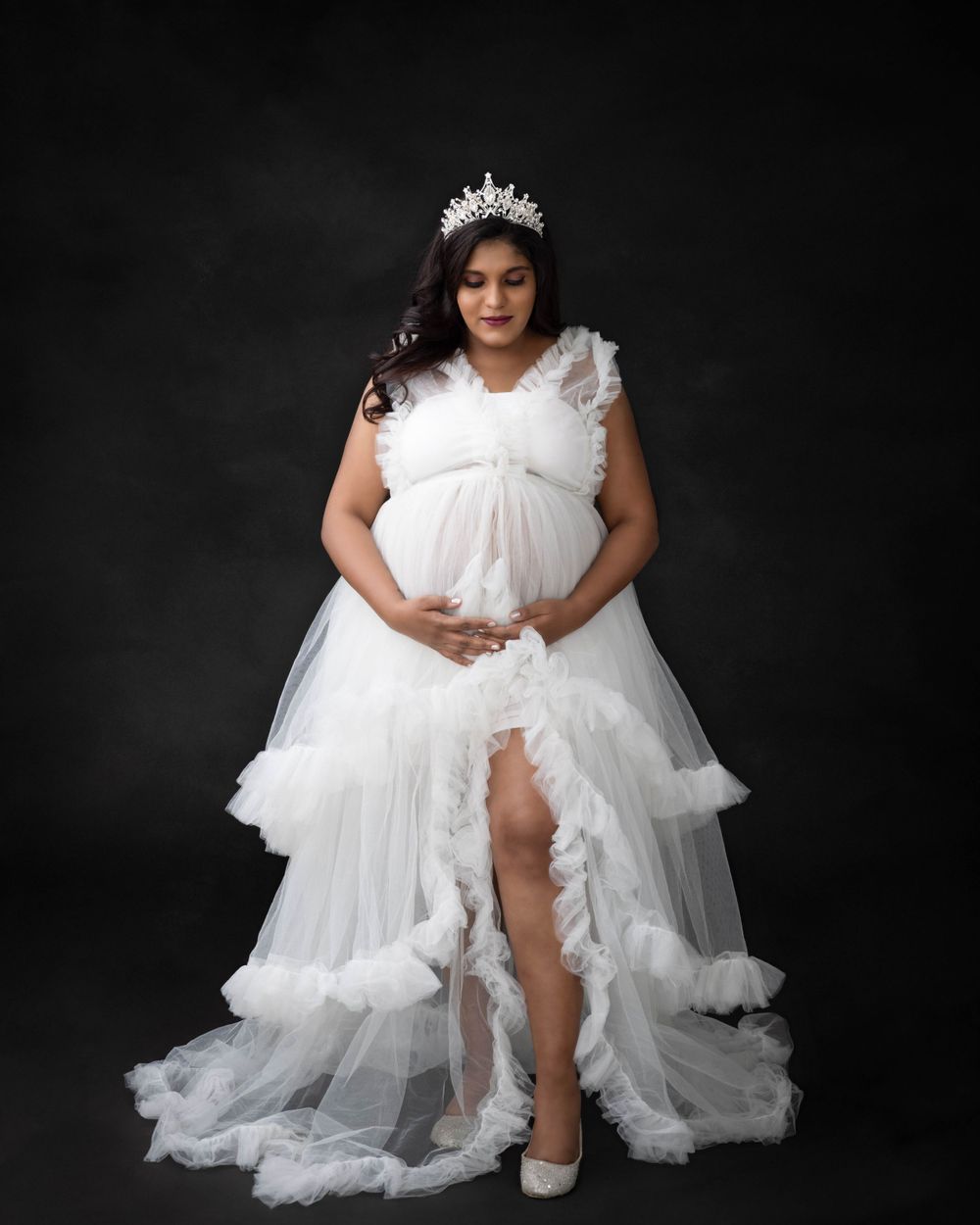 Elegant maternity photo in white tiered gown with crown accessory against dark backdrop.