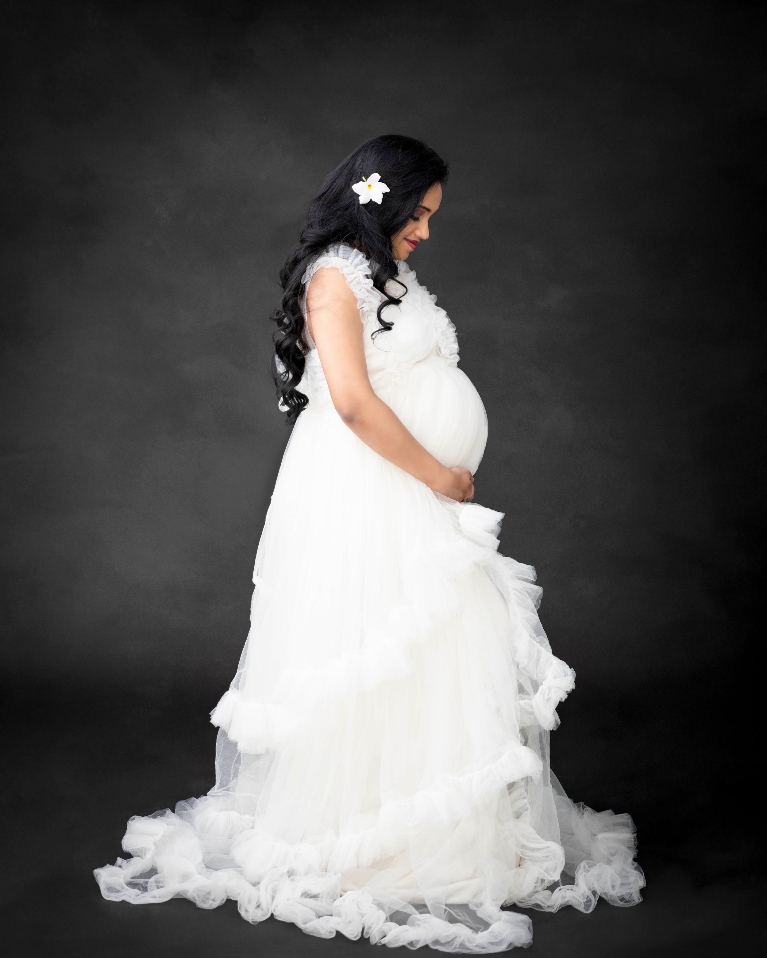 Side view maternity portrait in flowing white gown with floral hair accessory.