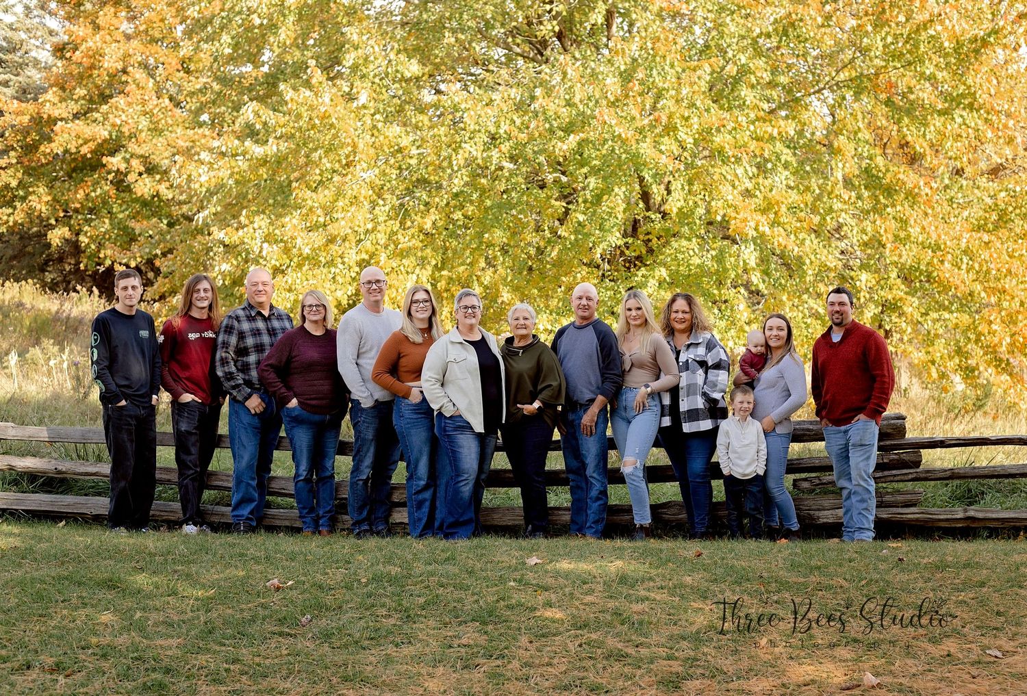 Extended family portrait group gathered outdoors in front of vibrant autumn foliage and wooden fence.