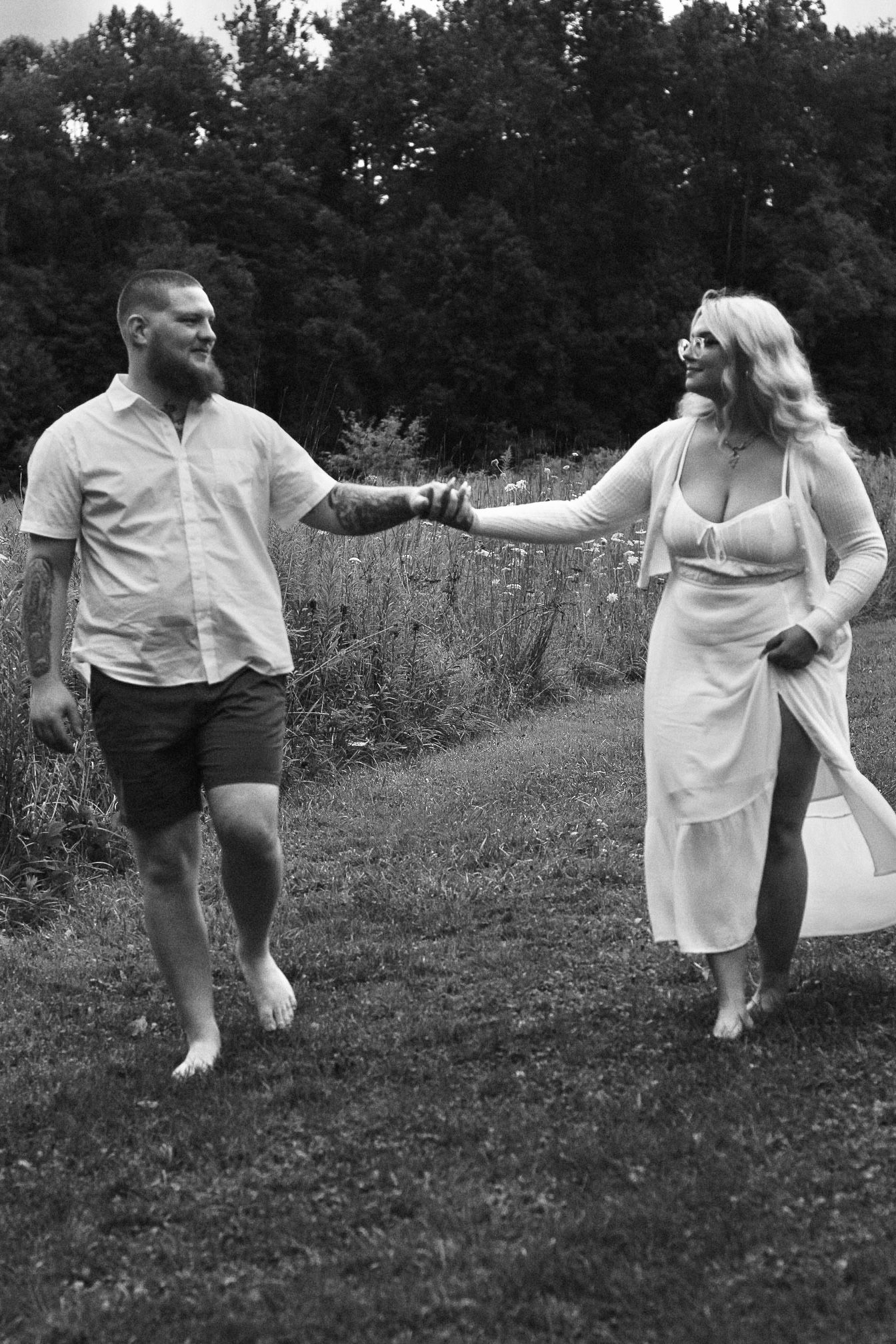 Black and white photo of couple playfully holding hands while walking through meadow.