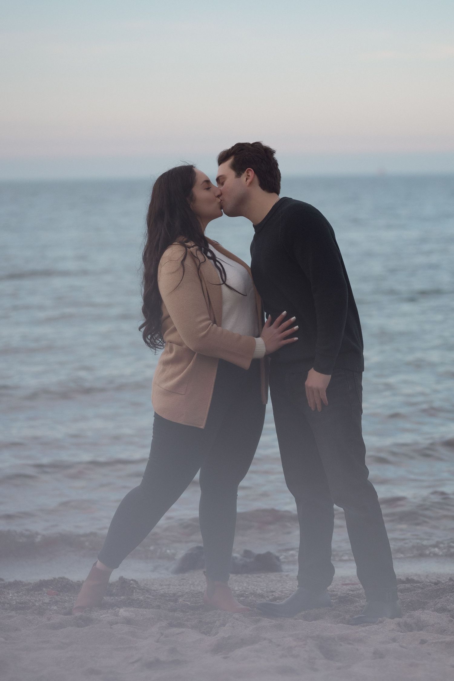 Couple sharing a tender kiss while standing barefoot on a foggy beach at sunset.
