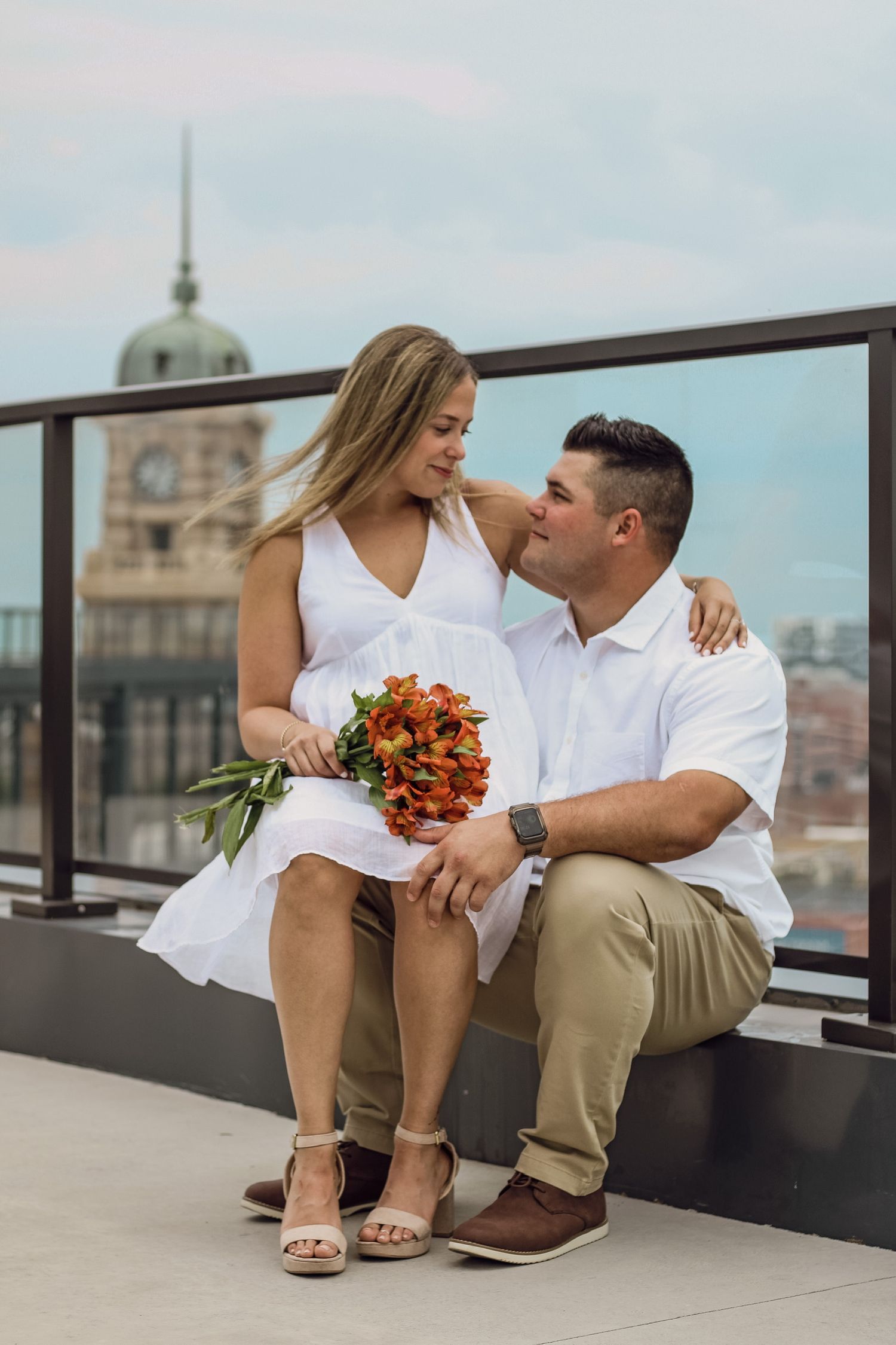 A couple sits on a rooftop holding an orange bouquet with city architecture behind them.