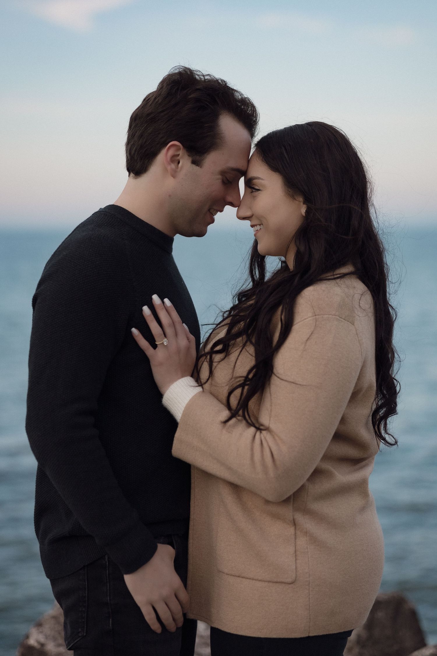Intimate engagement portrait of couple in sweaters against ocean backdrop.