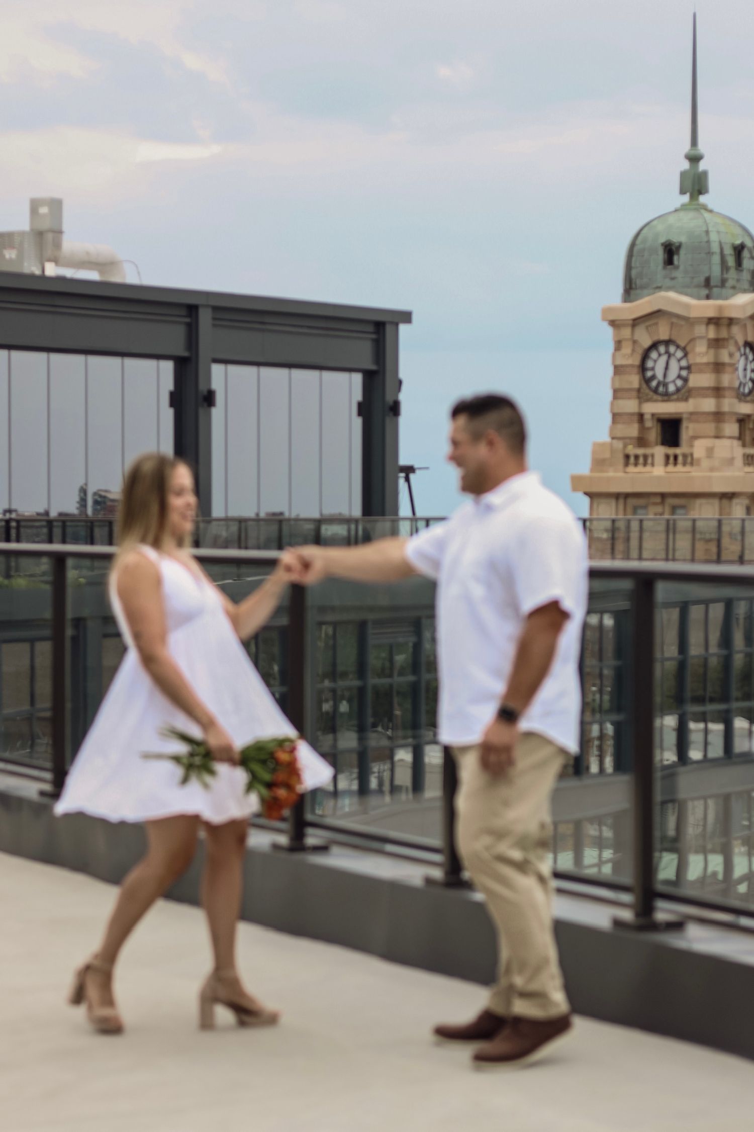A couple dances on a rooftop terrace with a historic clock tower in the background.