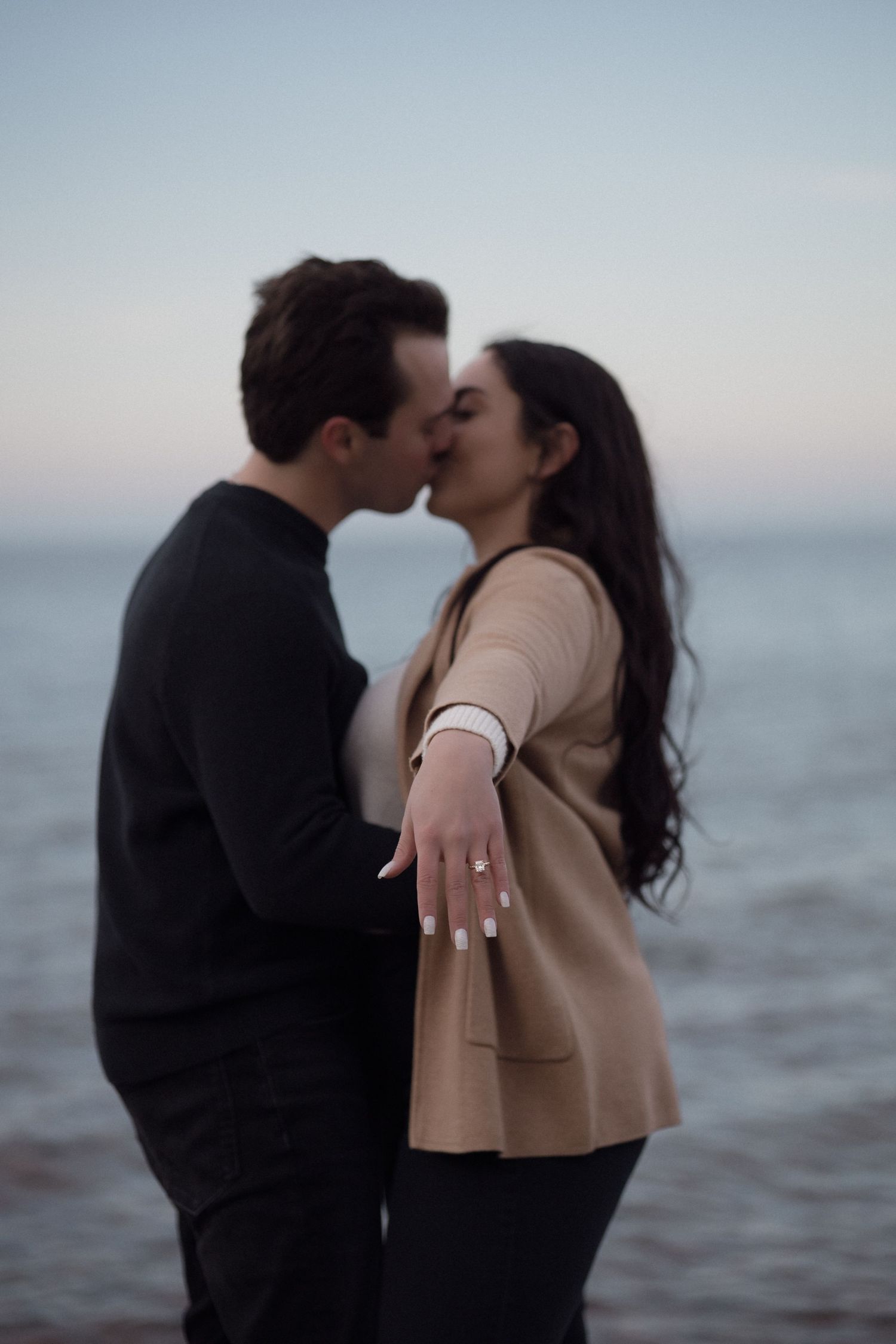 A romantic seaside engagement photo showing a couple kissing with an ocean backdrop at sunset.