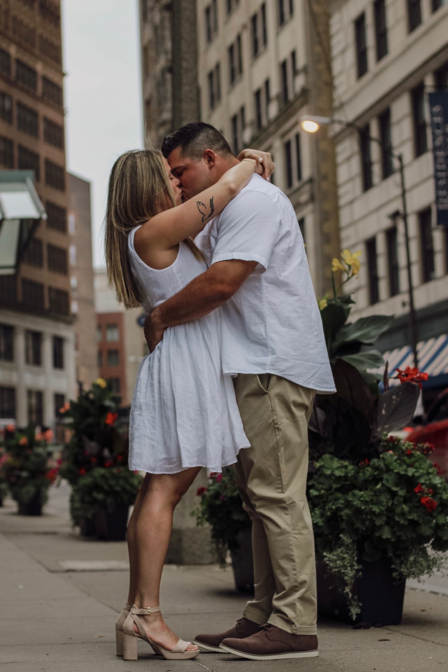 A couple shares an intimate moment on a city street lined with flowers and buildings.