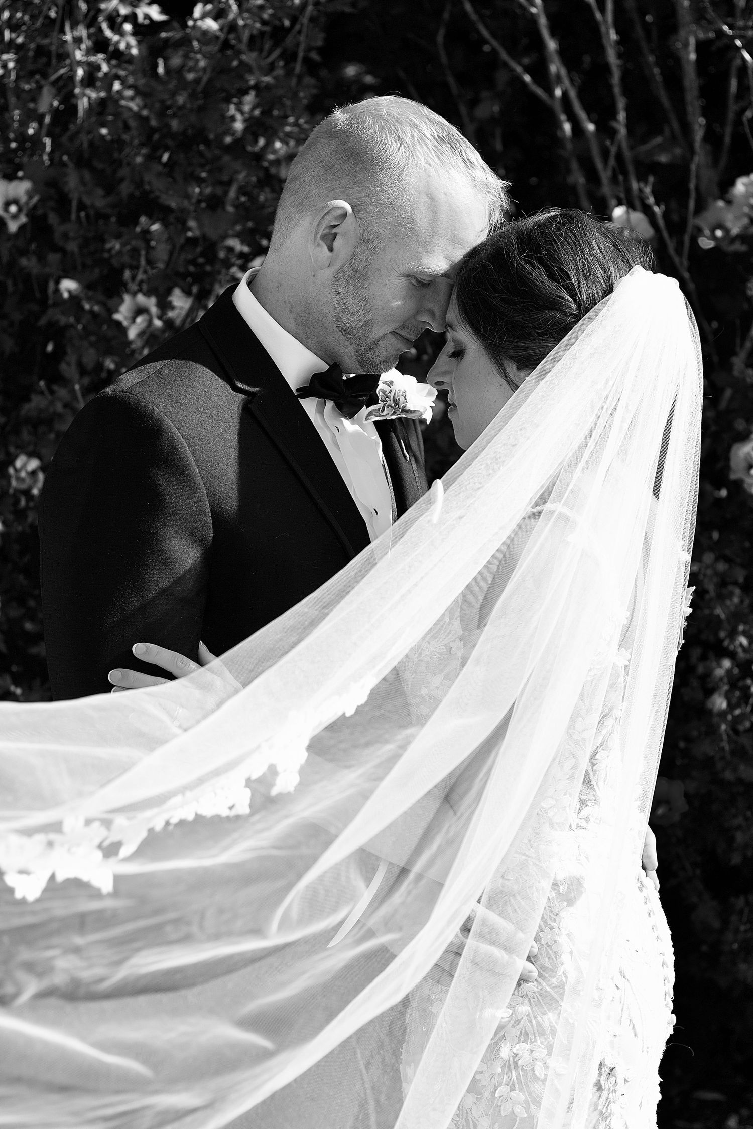 A dramatic black and white wedding photo showing a couple's intimate moment with flowing veil.