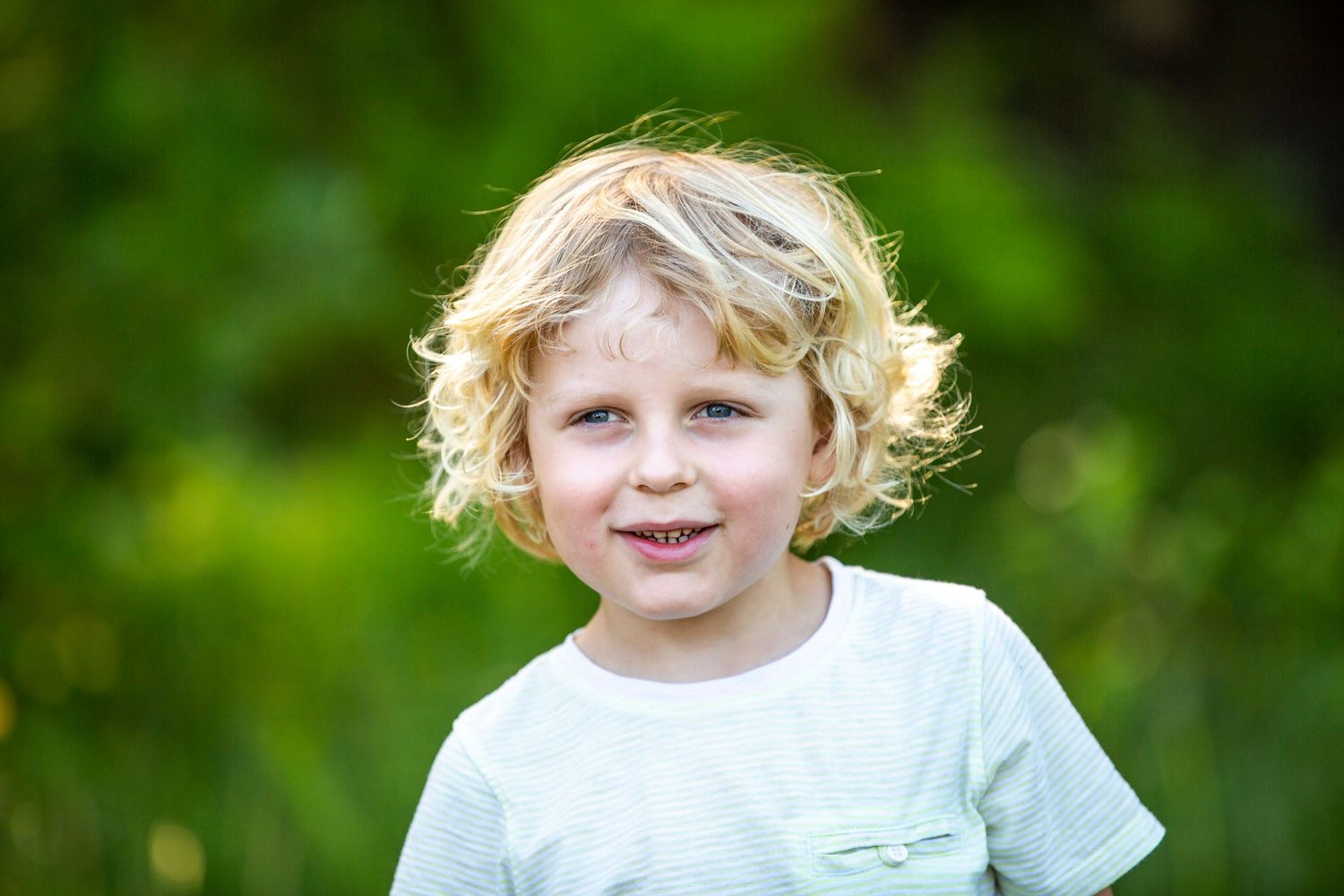 Young child with curly blonde hair smiles outdoors against green background.