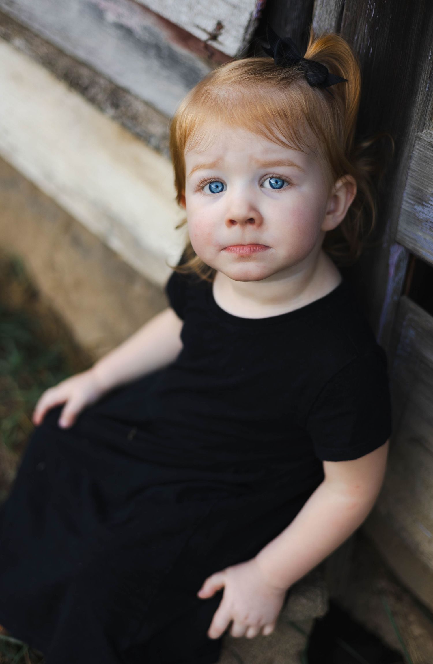 Young child in black dress sitting against rustic wall.
