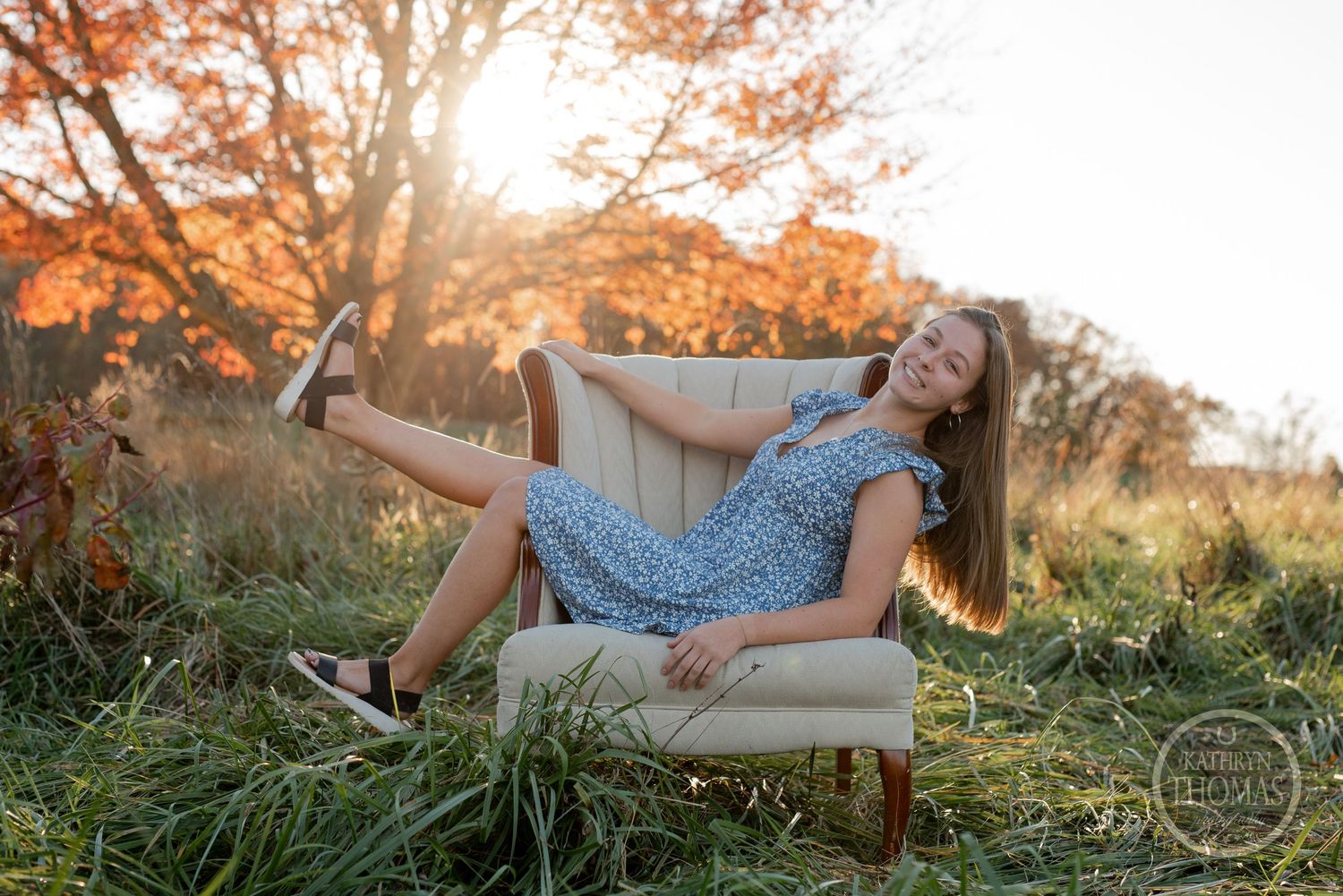 A casual pose on a beige armchair surrounded by tall grass and autumn trees at sunset.