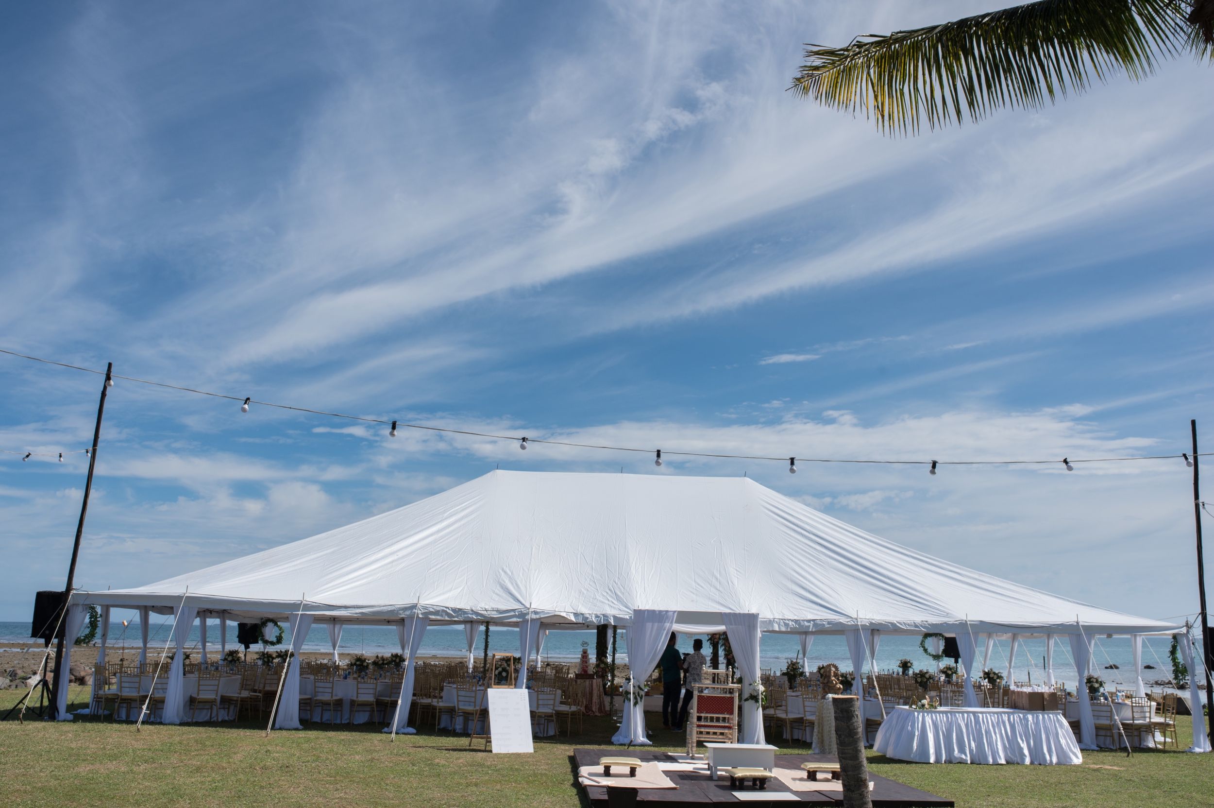 Large white wedding tent set up outdoors with string lights and palm trees against a blue sky.