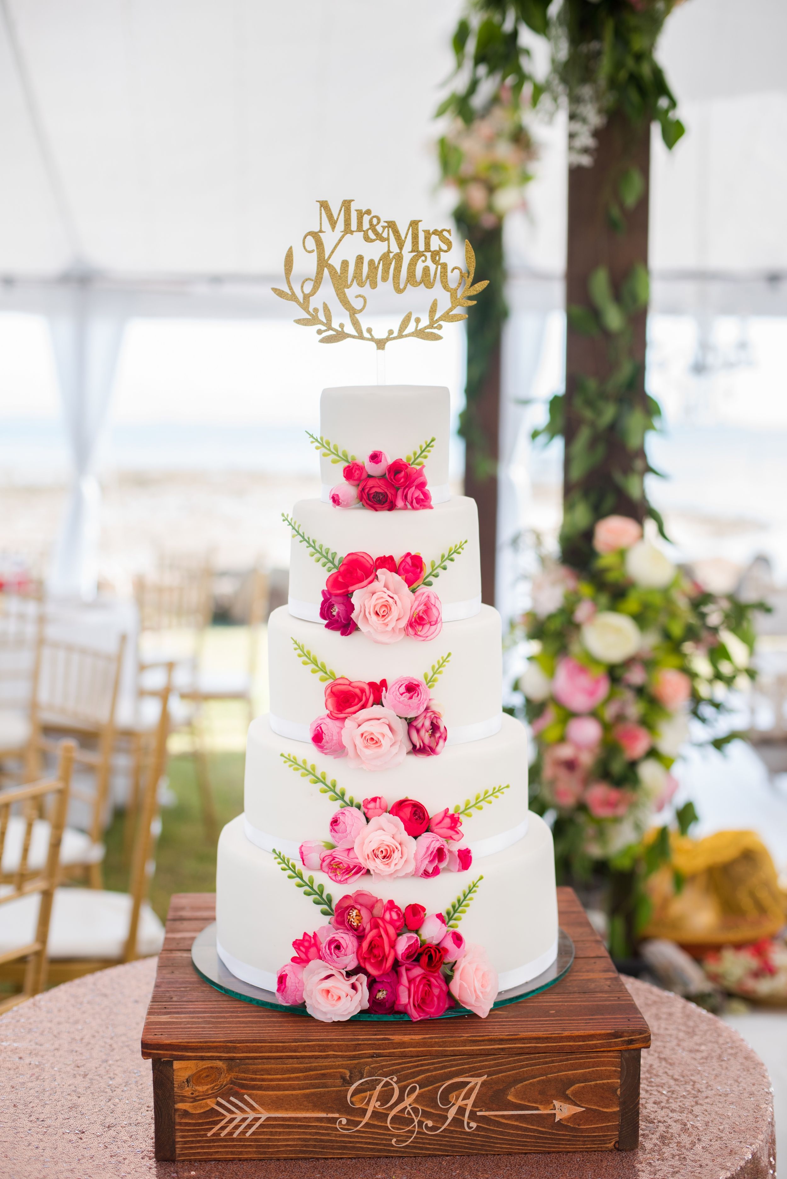 Four-tier white wedding cake decorated with pink roses and a gold 'Mrs & Mrs' topper.