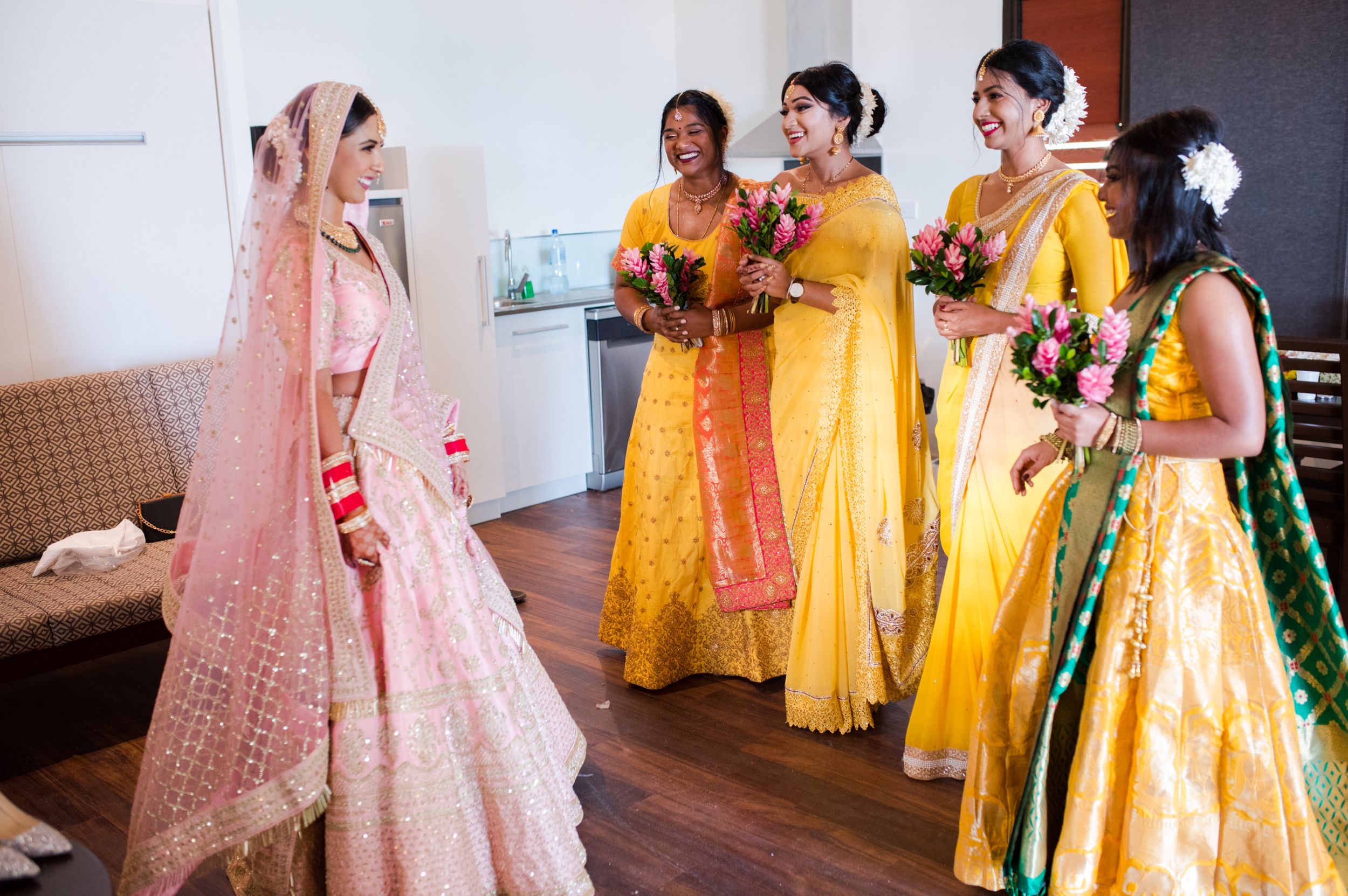 Bridesmaids in vibrant yellow lehengas smile and share a moment with bride in pink traditional attire during preparations.