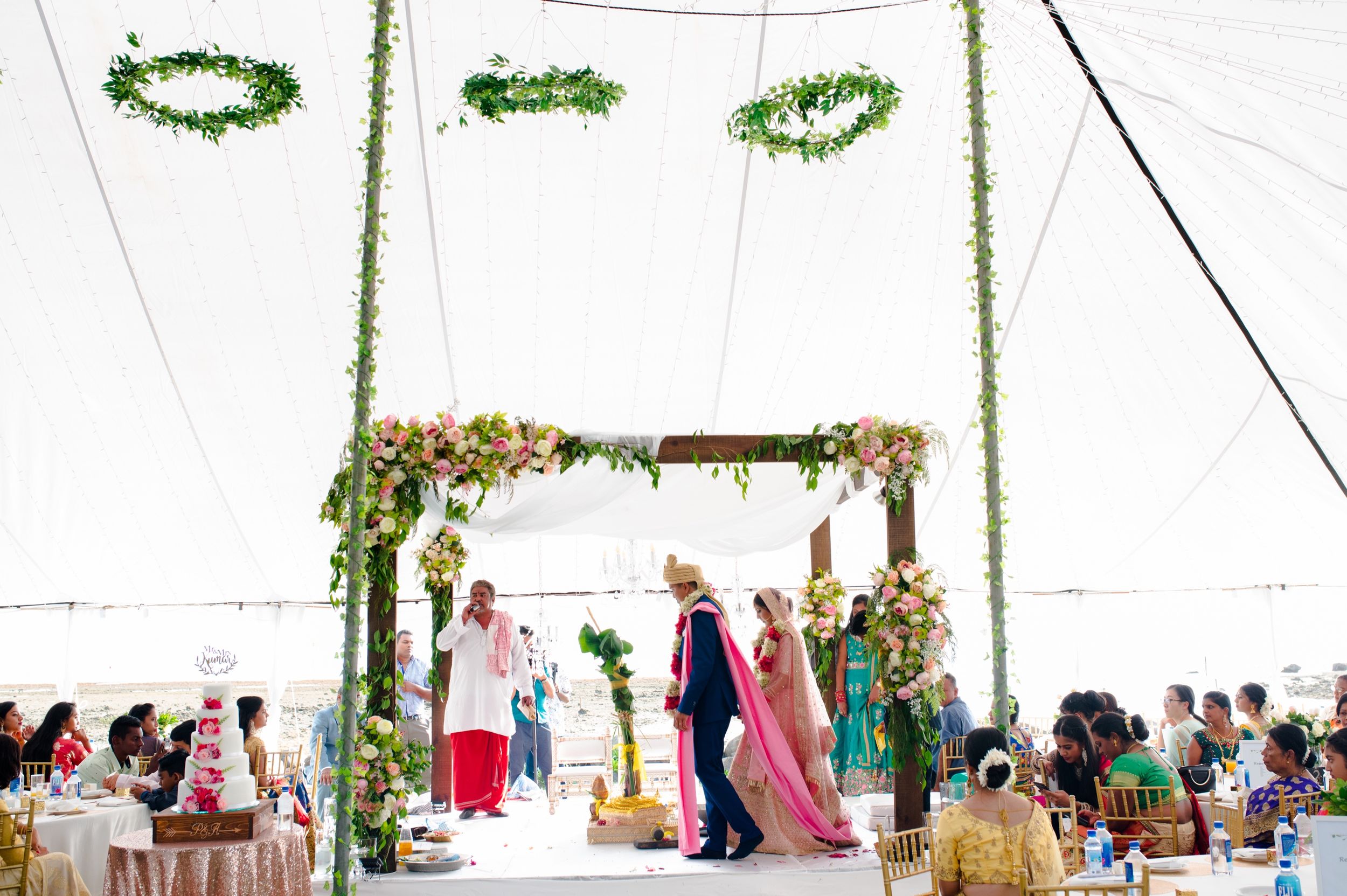 Wedding ceremony setup under a white tent with floral garlands and guests seated.