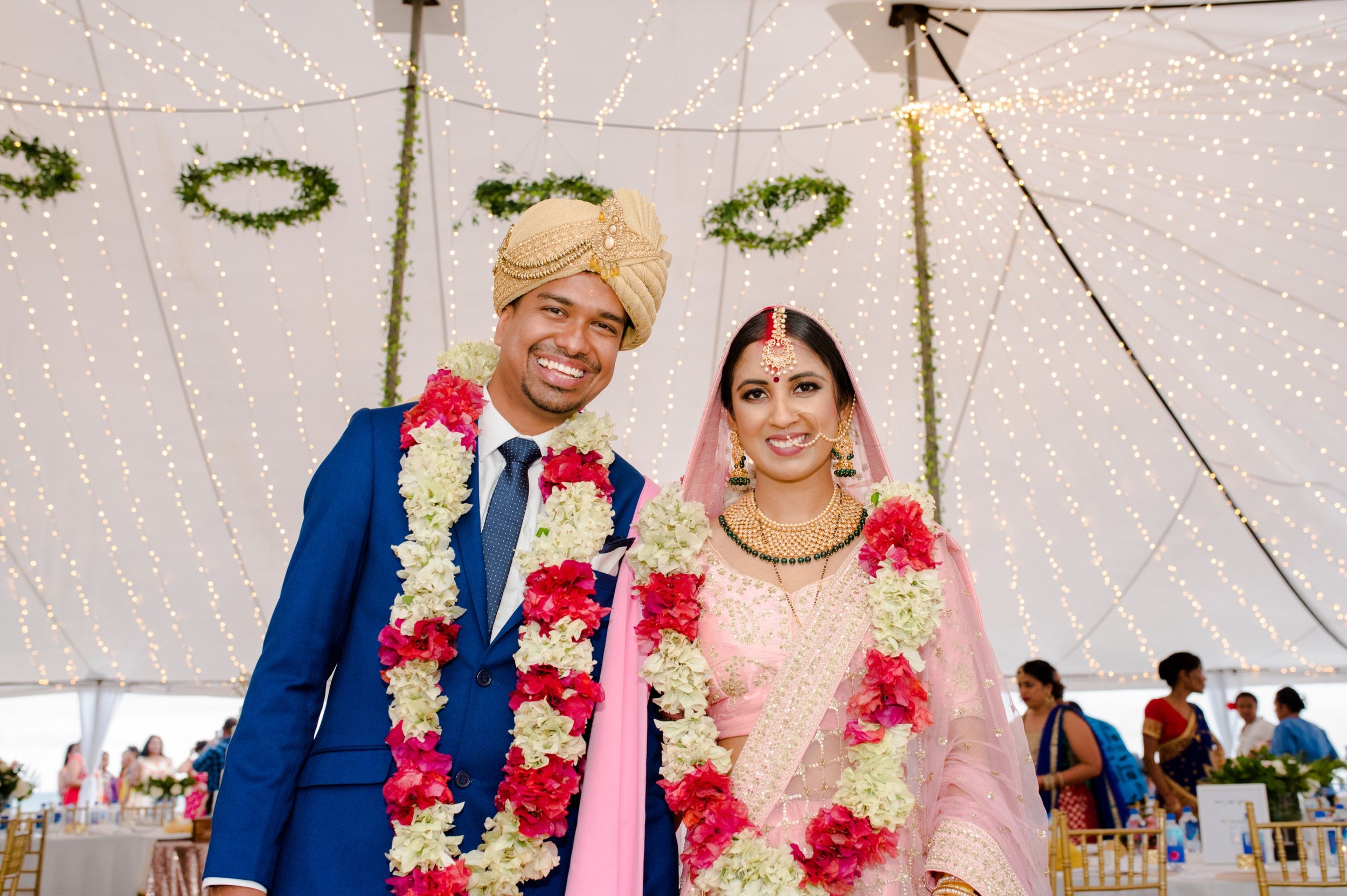Wedding couple stands together under a tent decorated with greenery and floral garlands in a traditional ceremony setting.