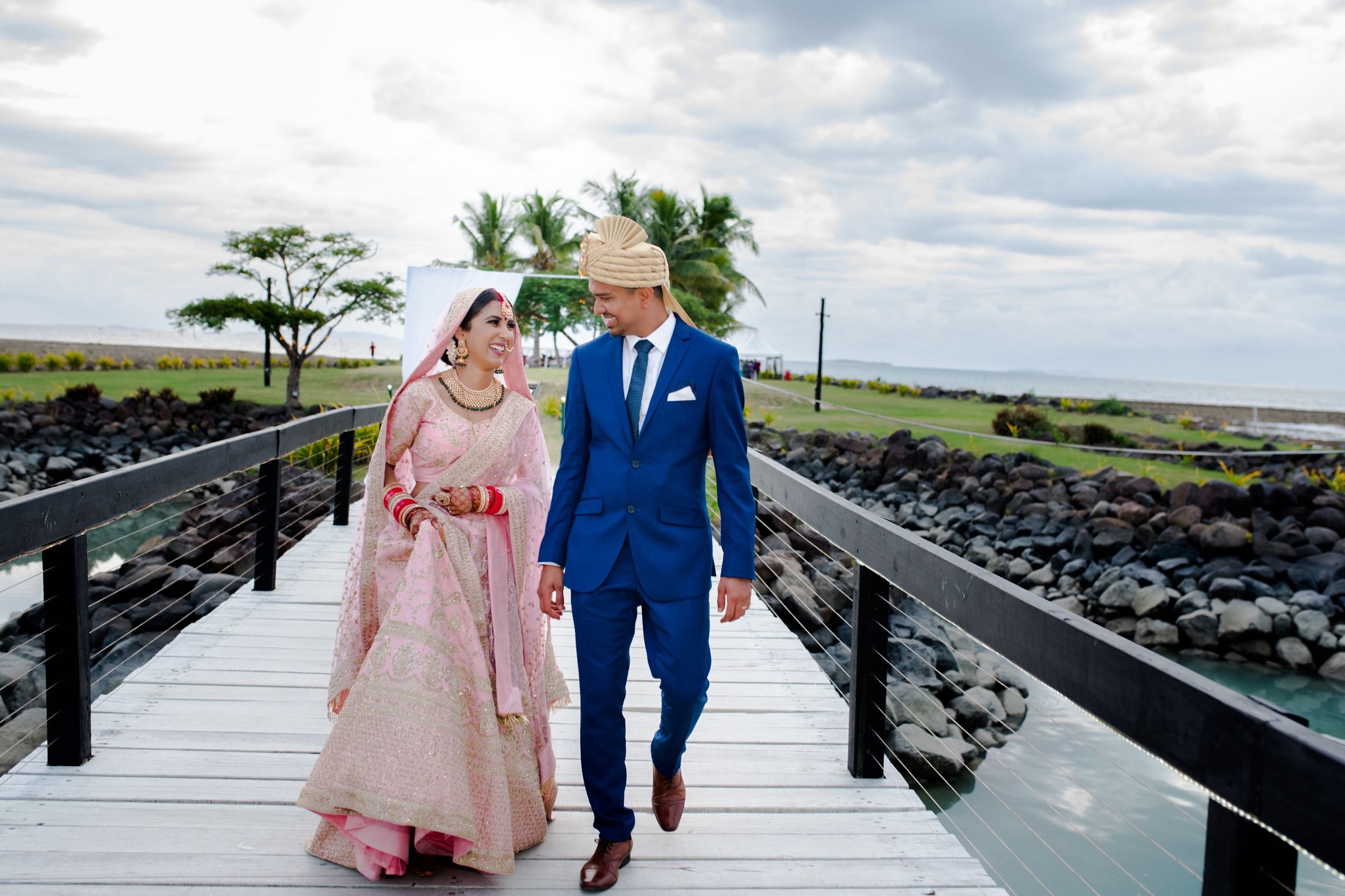 Couple in wedding attire walks along an oceanside boardwalk with palm trees and rocky coast in background.