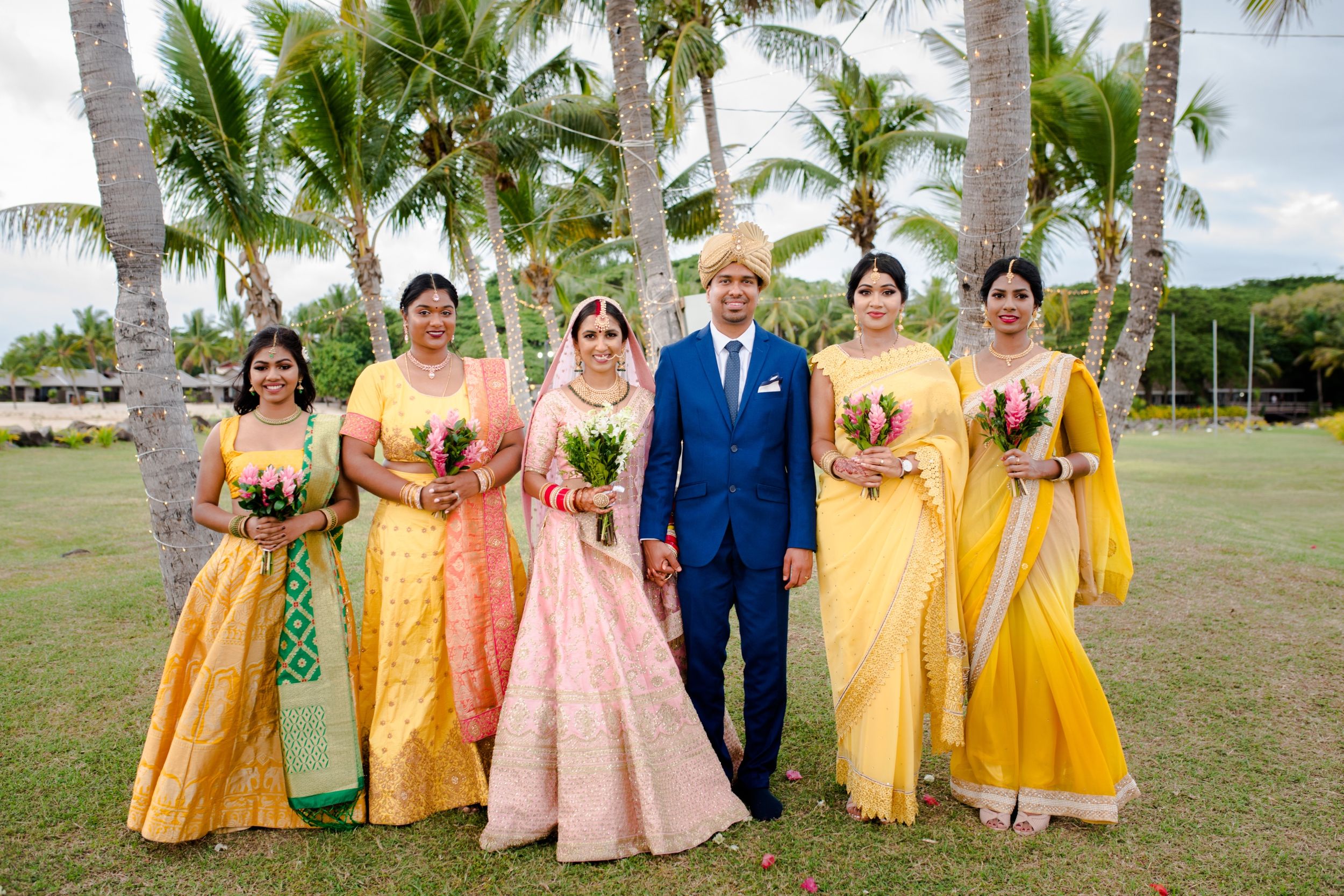 Wedding party in coordinated yellow and pink attire poses together under tropical palm trees.