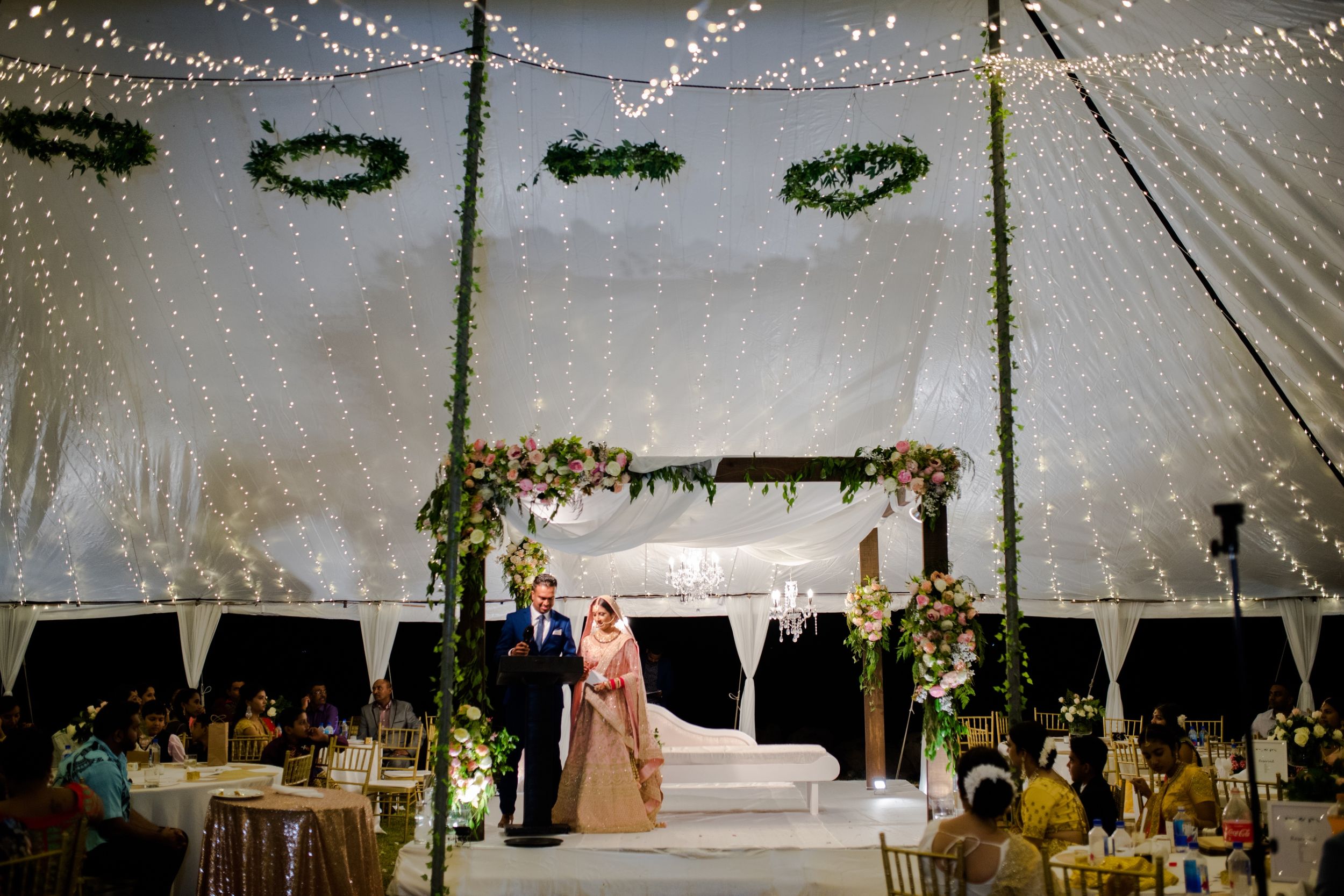 A beautifully decorated wedding tent with string lights and green wreaths illuminating the evening celebration.