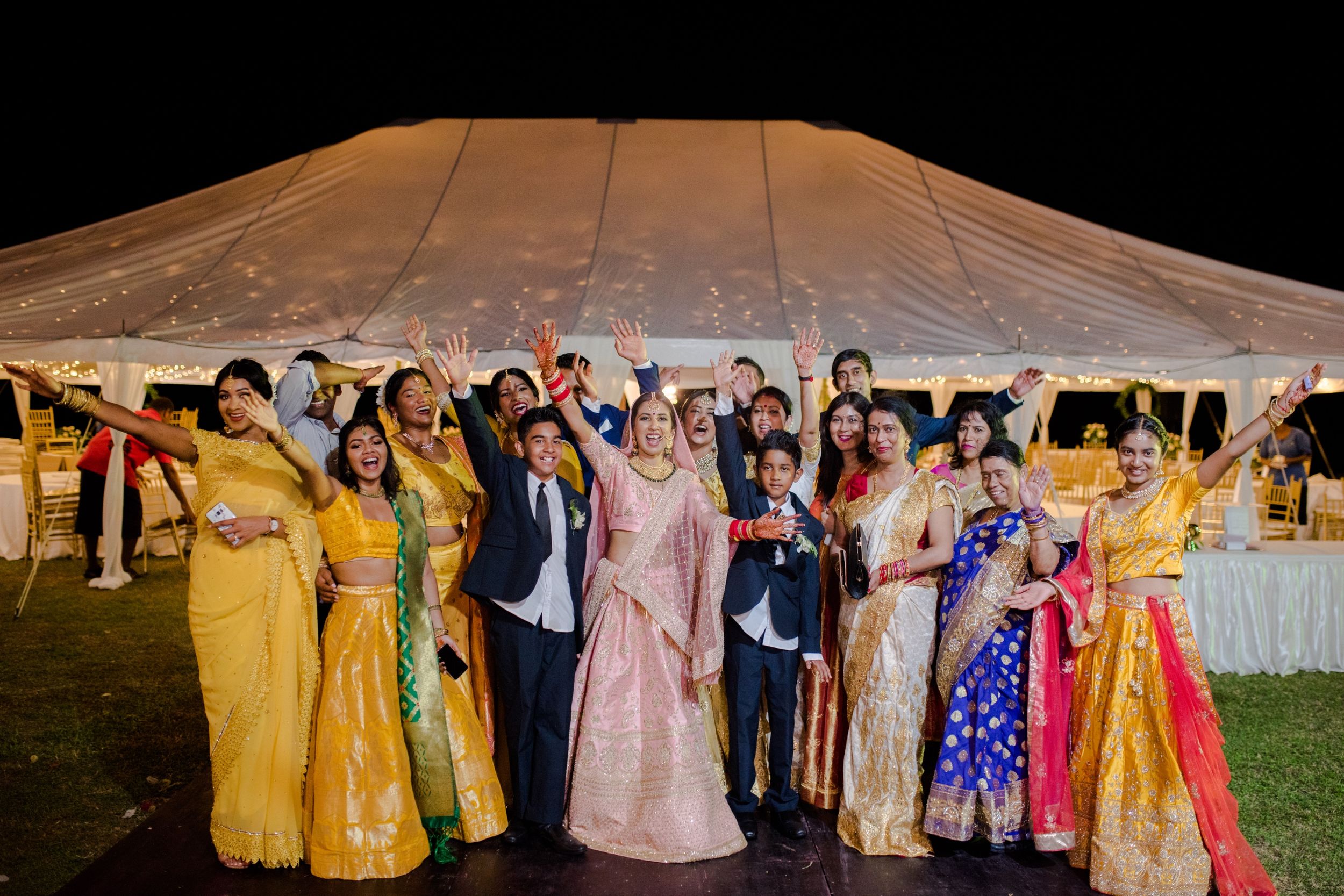 Group photo of wedding guests in vibrant traditional attire under a large white tent.