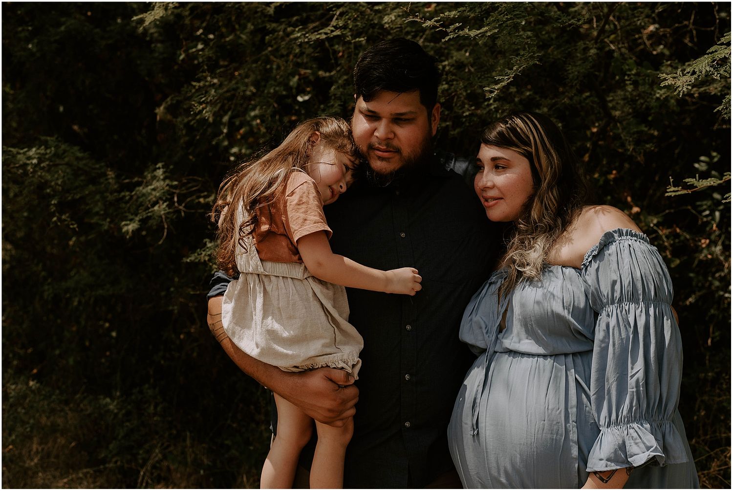 Family maternity portrait in woods featuring blue dress and peach-colored outfit.