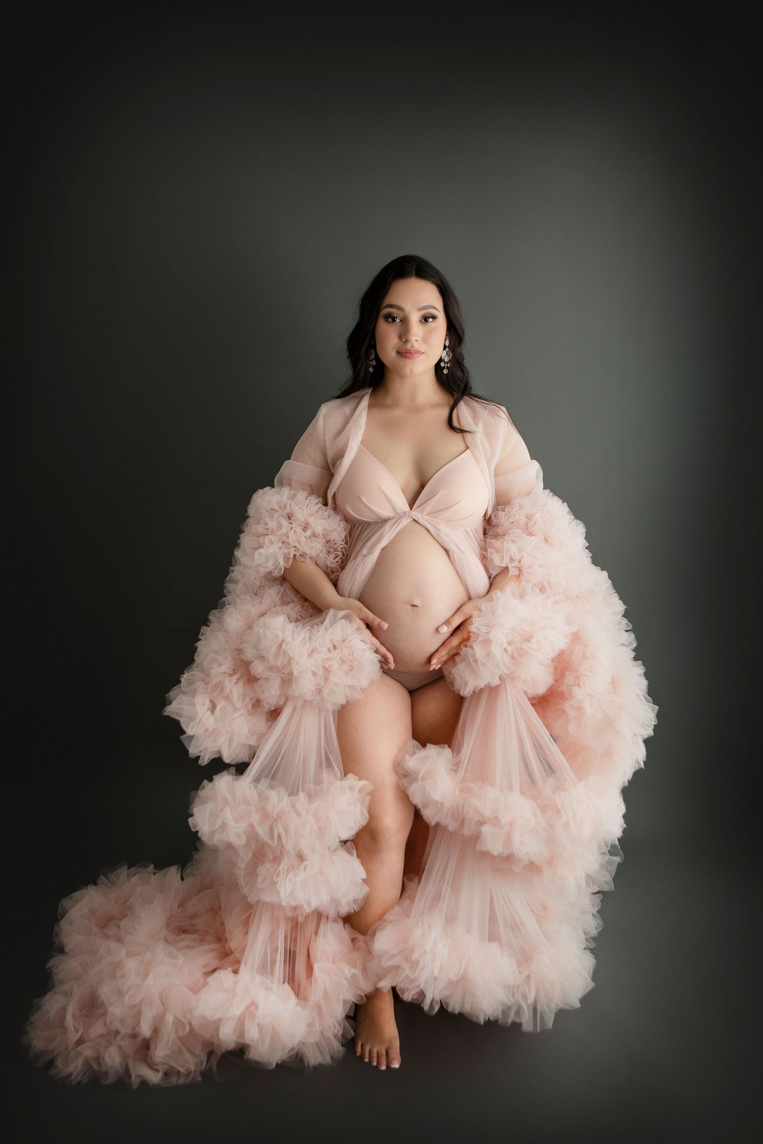 Maternity photo of woman wearing flowing pink tulle robe against dark background.