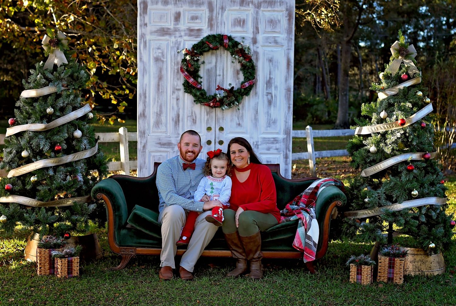 Holiday portrait setup with decorative white door, wreath, Christmas trees, and wrapped presents in an outdoor setting.