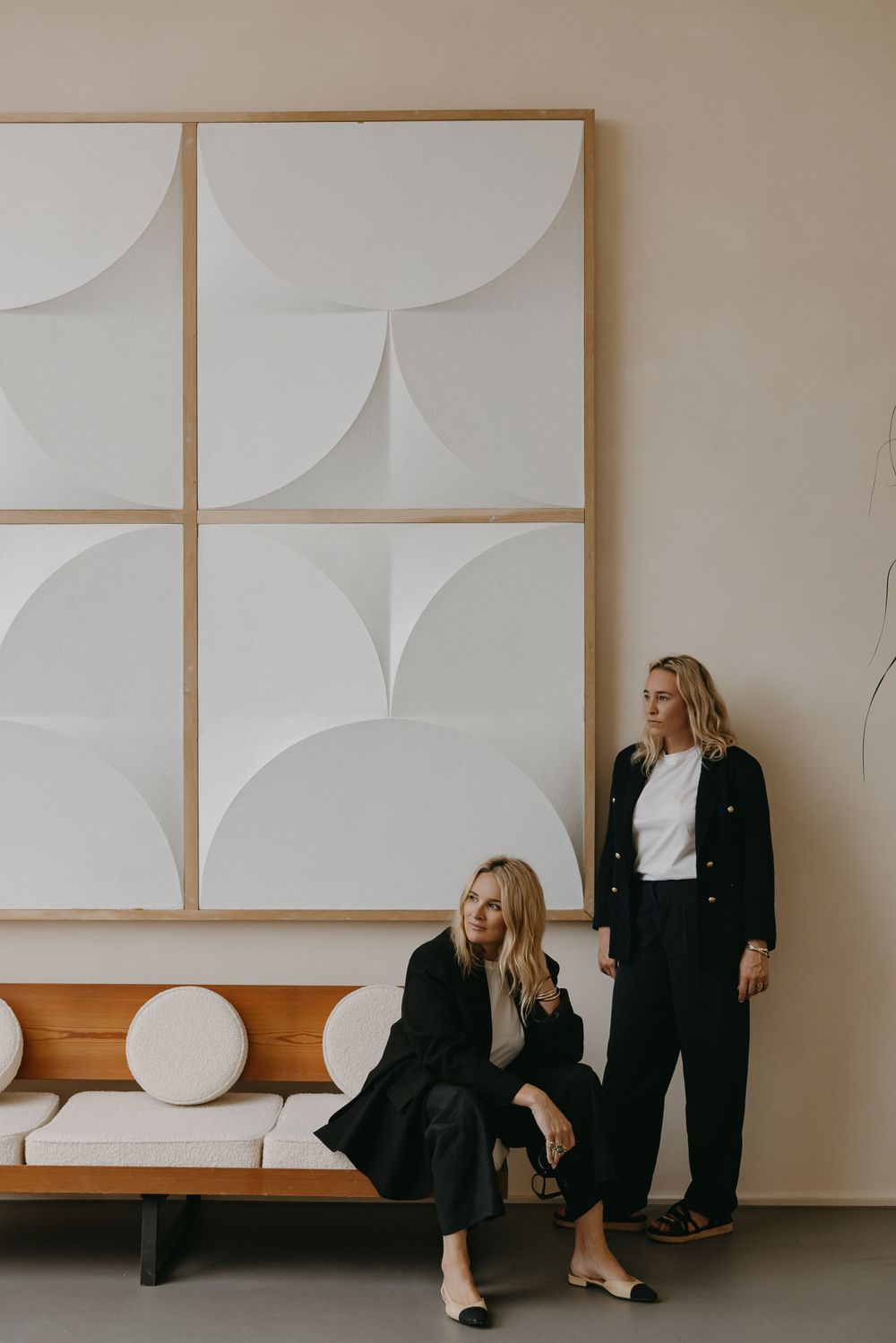 Two people in black outfits posing against minimalist white wall art and wooden bench.