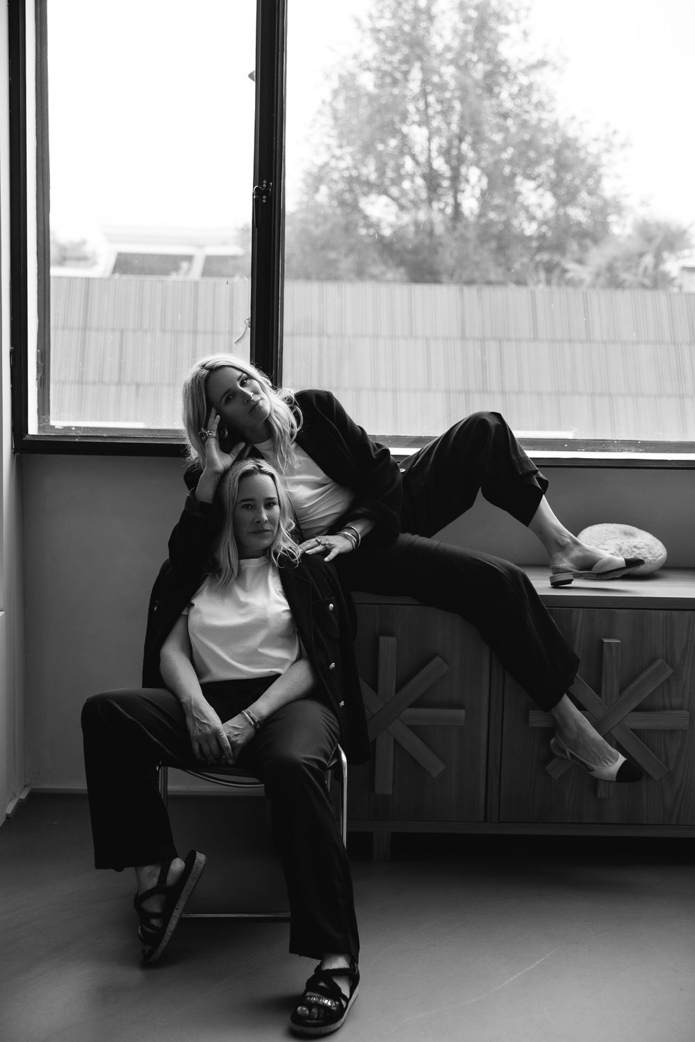 Two women in casual attire lounging by a window in black and white photography.