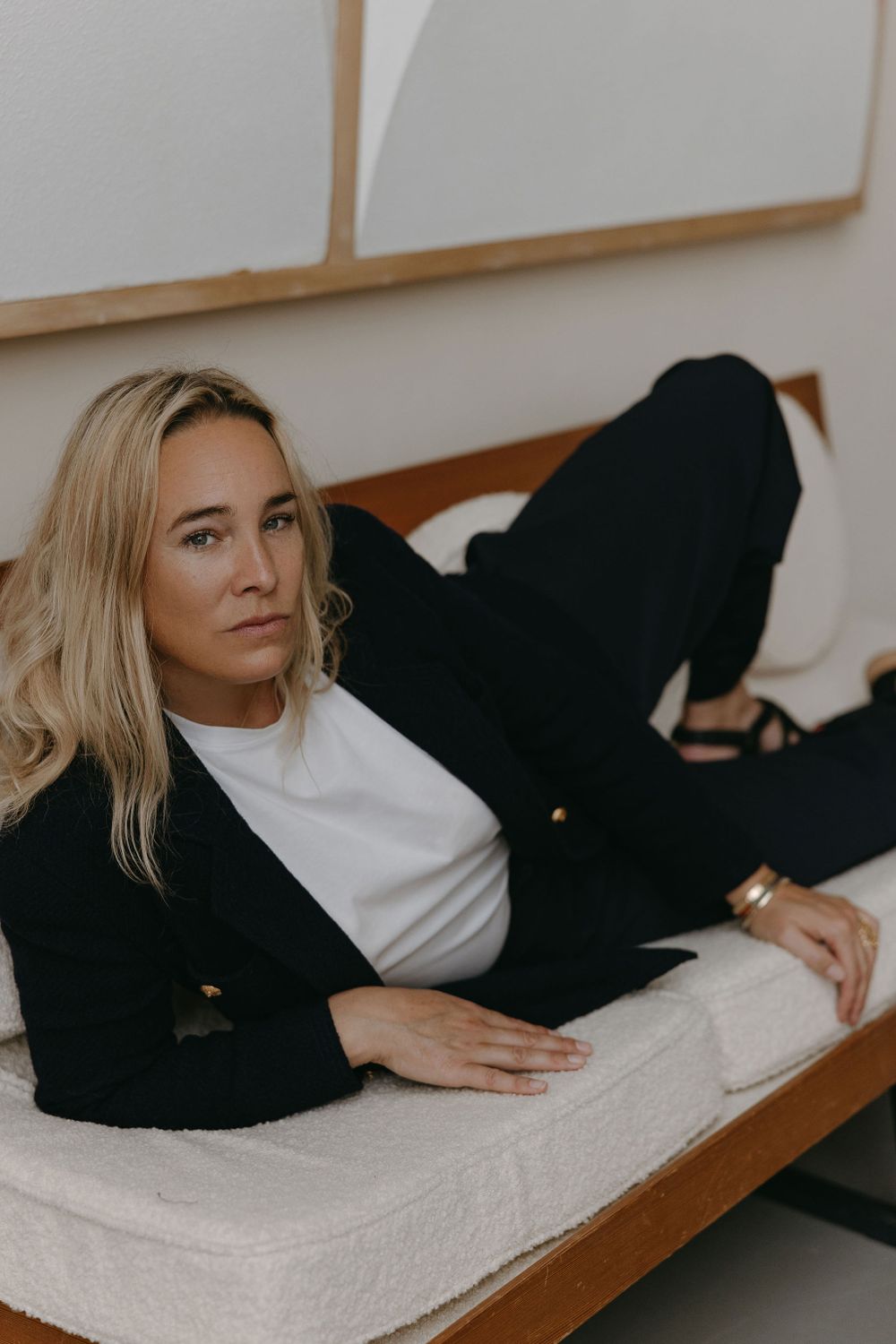 Woman in black blazer and white shirt lounging on bench against modern wall art.