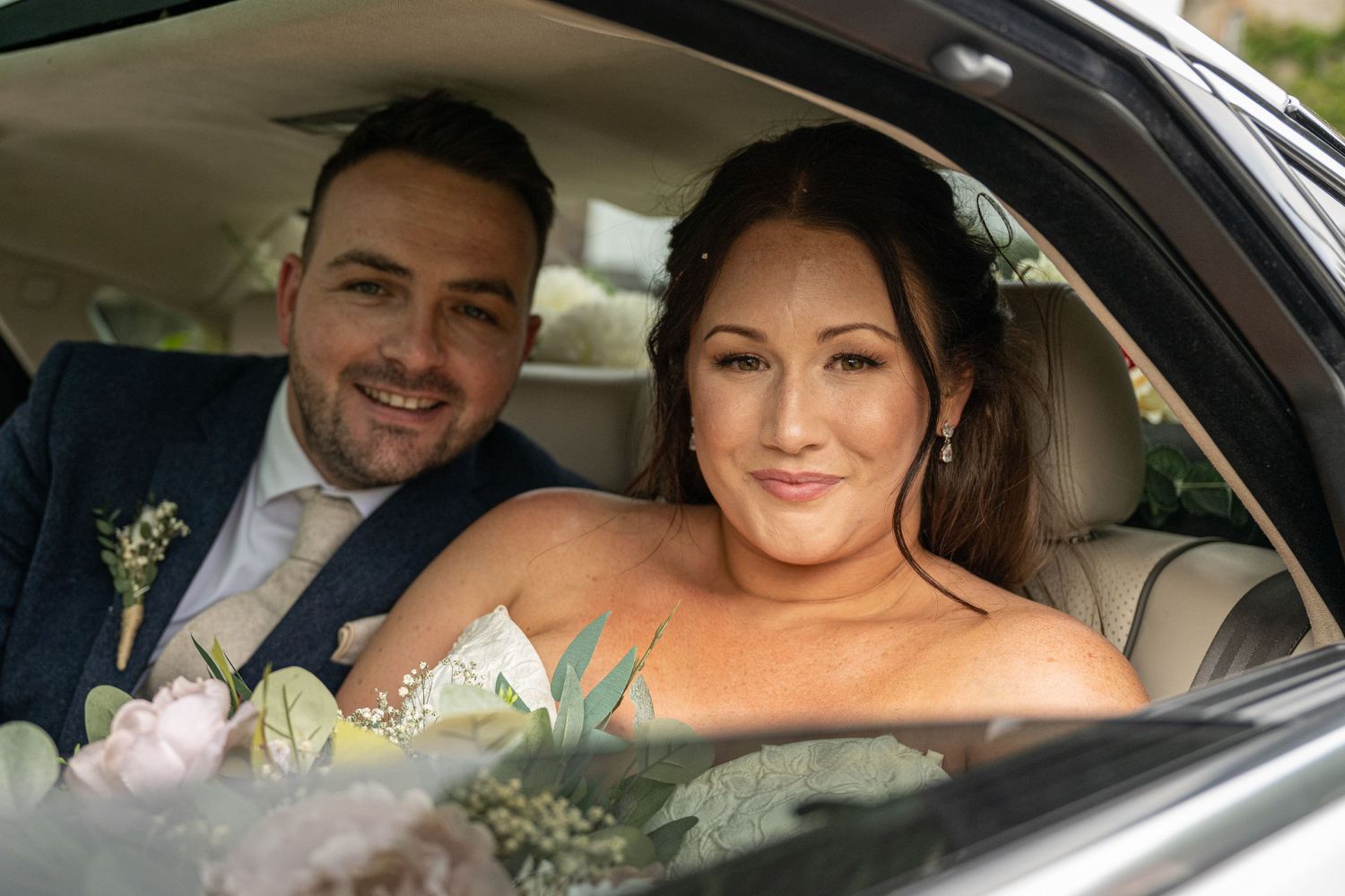 A couple smiles together in a car with wedding flowers visible.