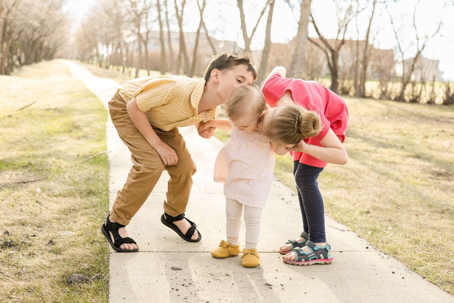 Three people bend down to play together on a sunny path in a park with bare trees in the background.