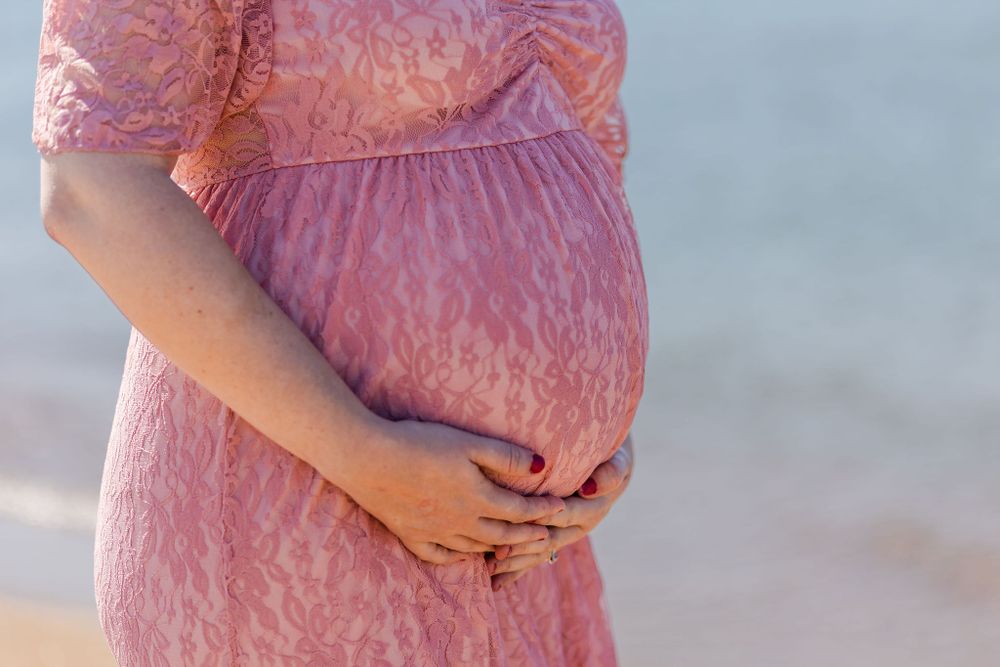 Close up of pregnant belly in pink lace dress with hands gently cradling bump against ocean backdrop.