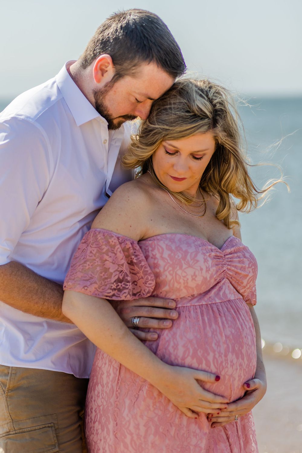 Romantic maternity photo on the beach with couple embracing in pink dress and white shirt at sunset.