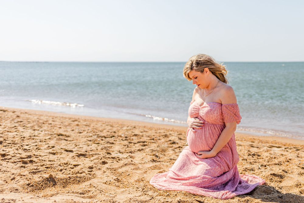 Person in pink dress sitting on beach during sunset maternity photo session.