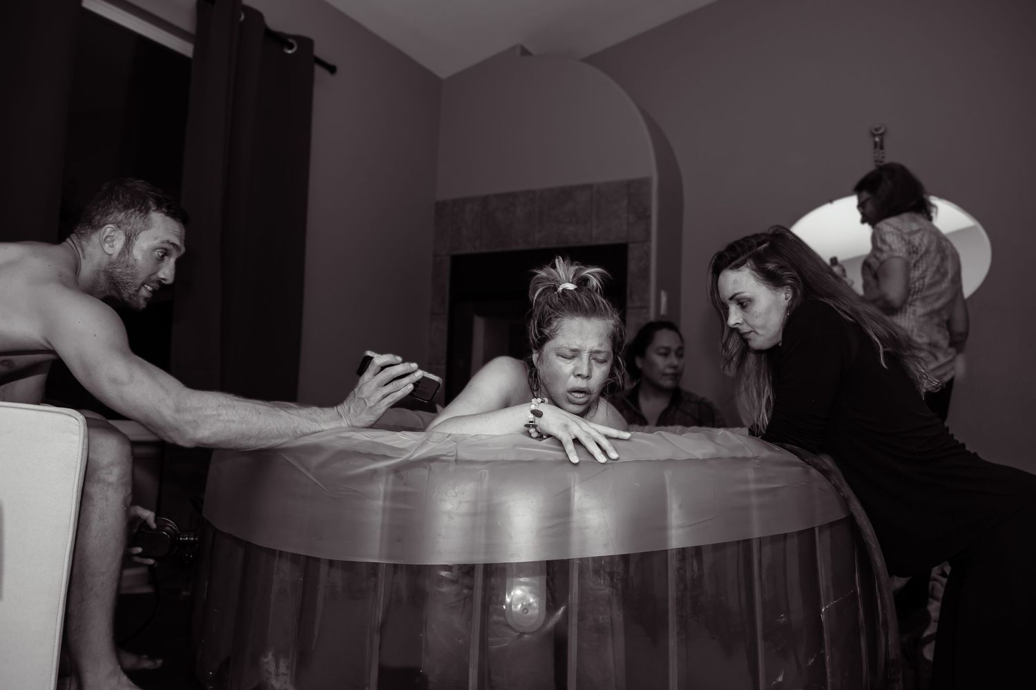 Black and white image of a pregnant woman in labor in a birthing tub in her home, surrounded by her support people.