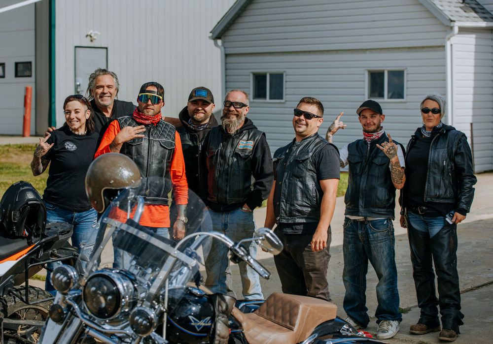 Group of motorcycle enthusiasts in leather gear gathered around bikes outside white building.
