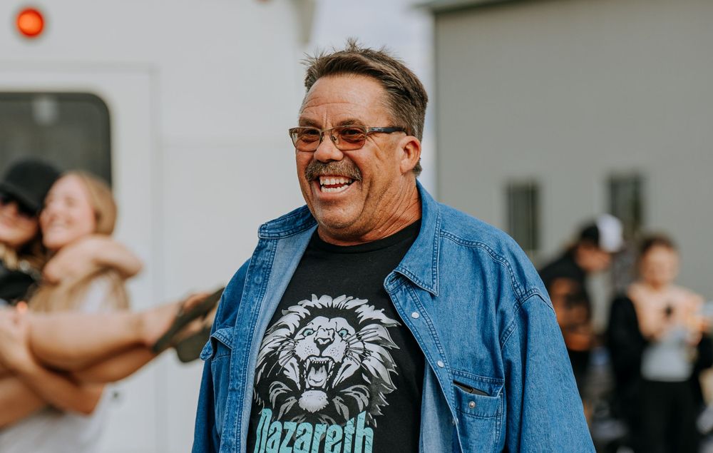 A person wearing a denim jacket with a lion graphic tee smiles warmly outdoors.